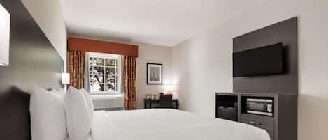 A modern hotel room with a large bed, a flat-screen TV mounted on the wall, a desk with a lamp, and a window with curtains.