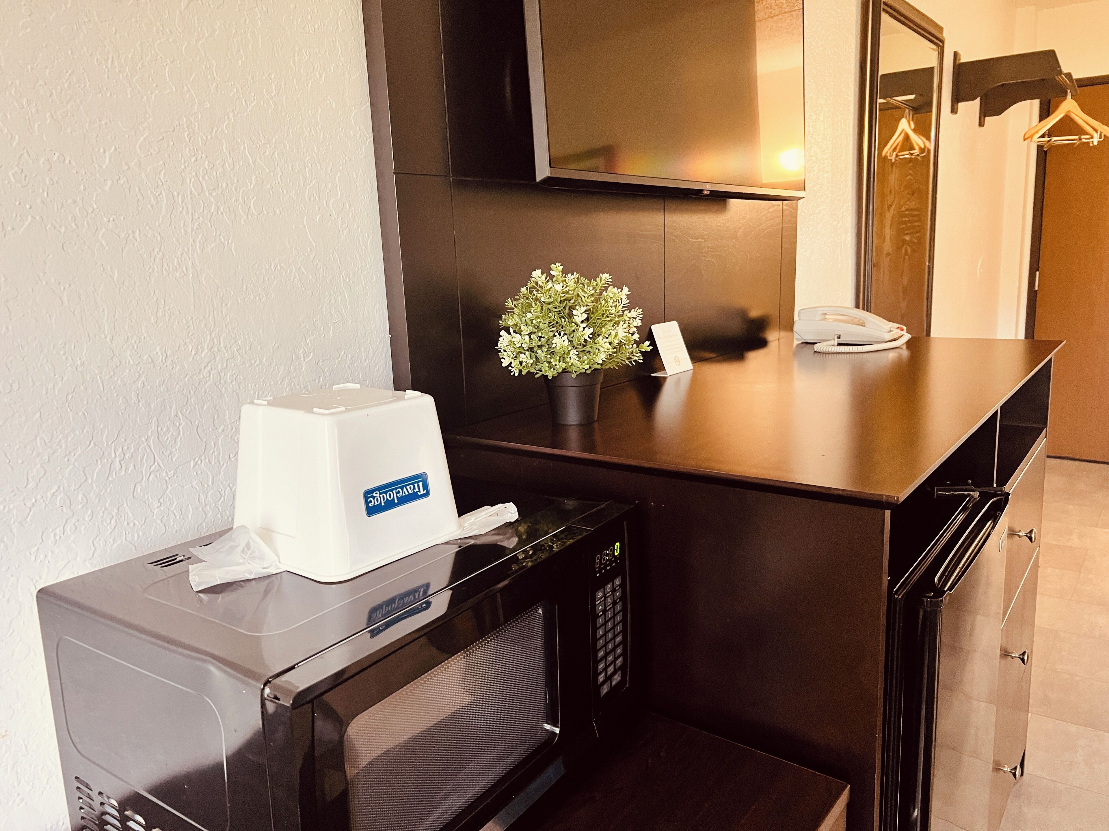 Een compacte kitchenette met een magnetron, een kleine koelkast, een prullenbak, een bloempot en een telefoon op het aanrecht.