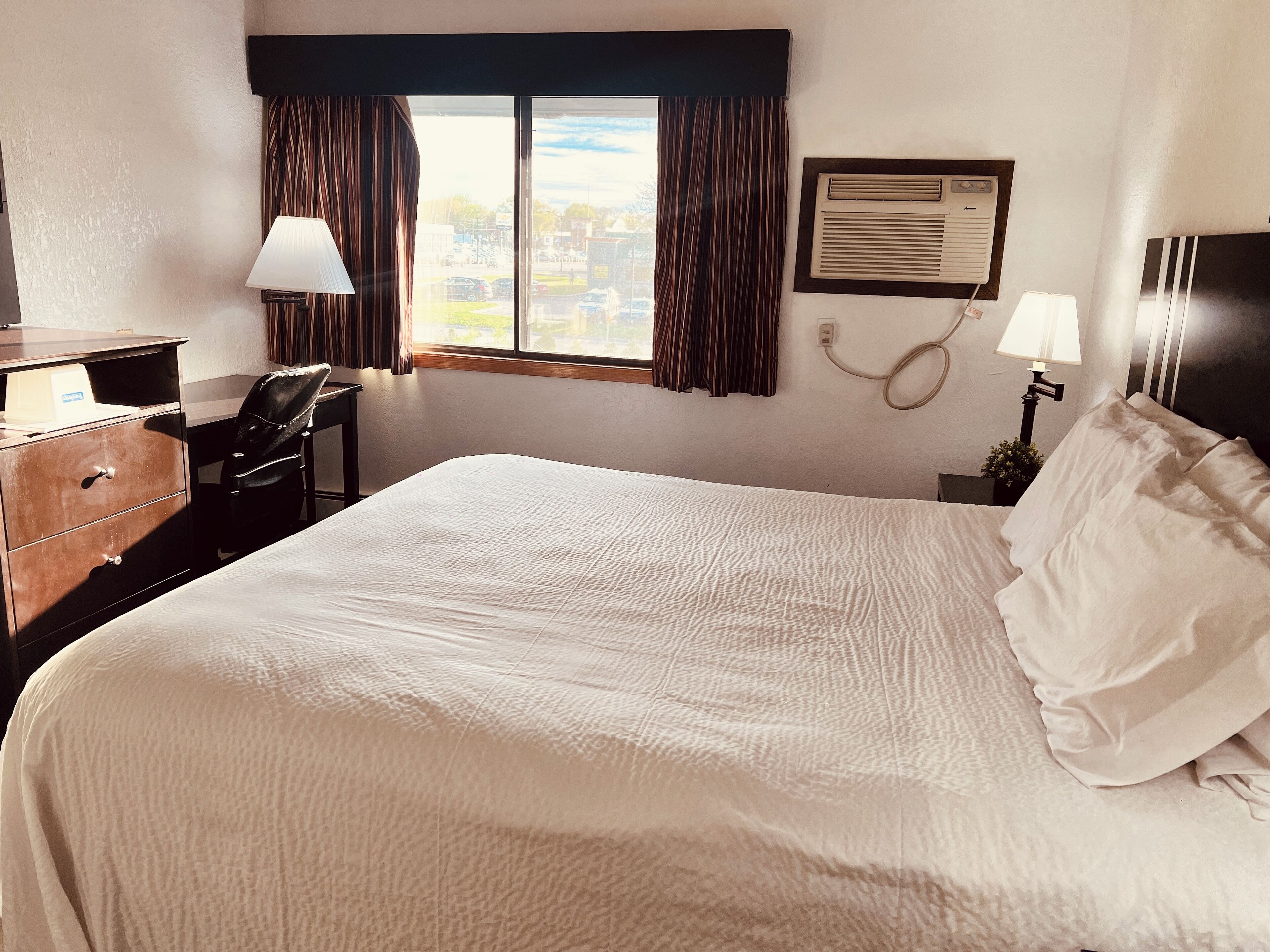 A hotel room with a large bed, a desk with a chair, a lamp, and a window with curtains.