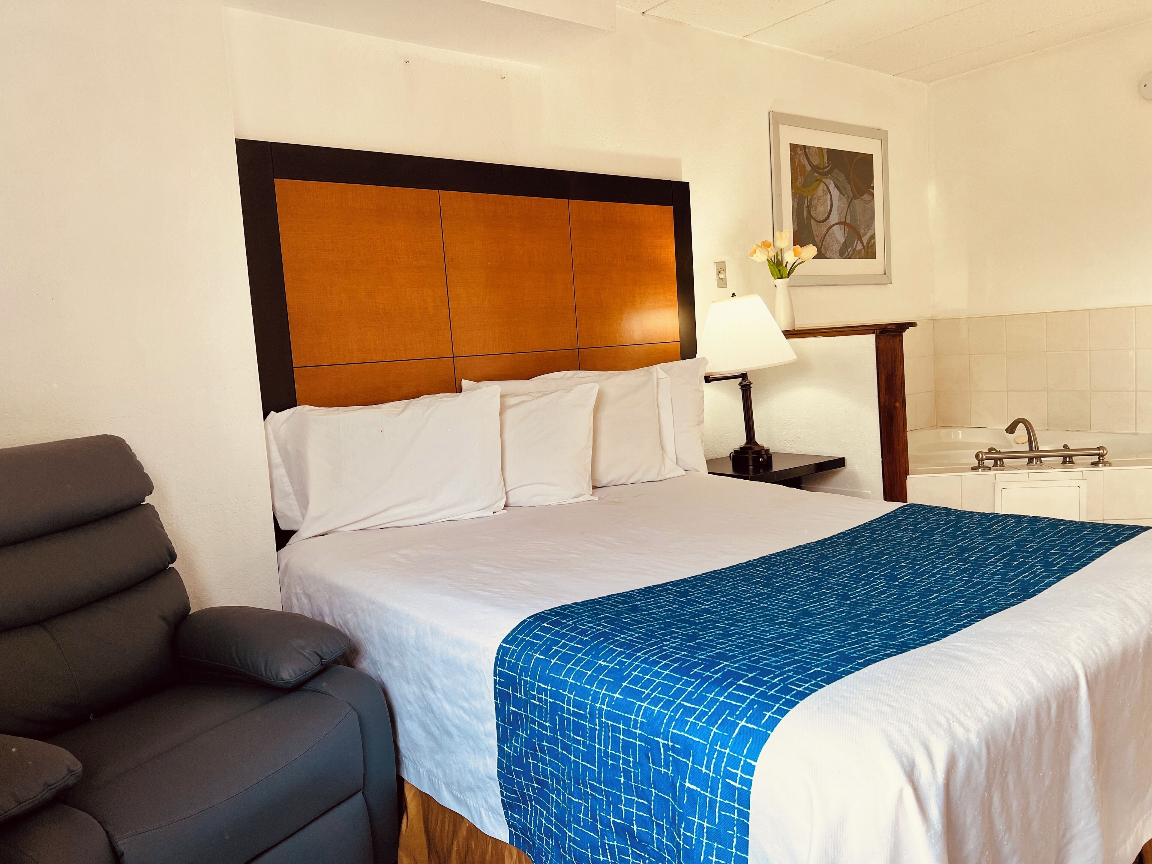 A hotel room with a bed, a nightstand, a lamp, a chair, and a bathroom with a bathtub and sink.