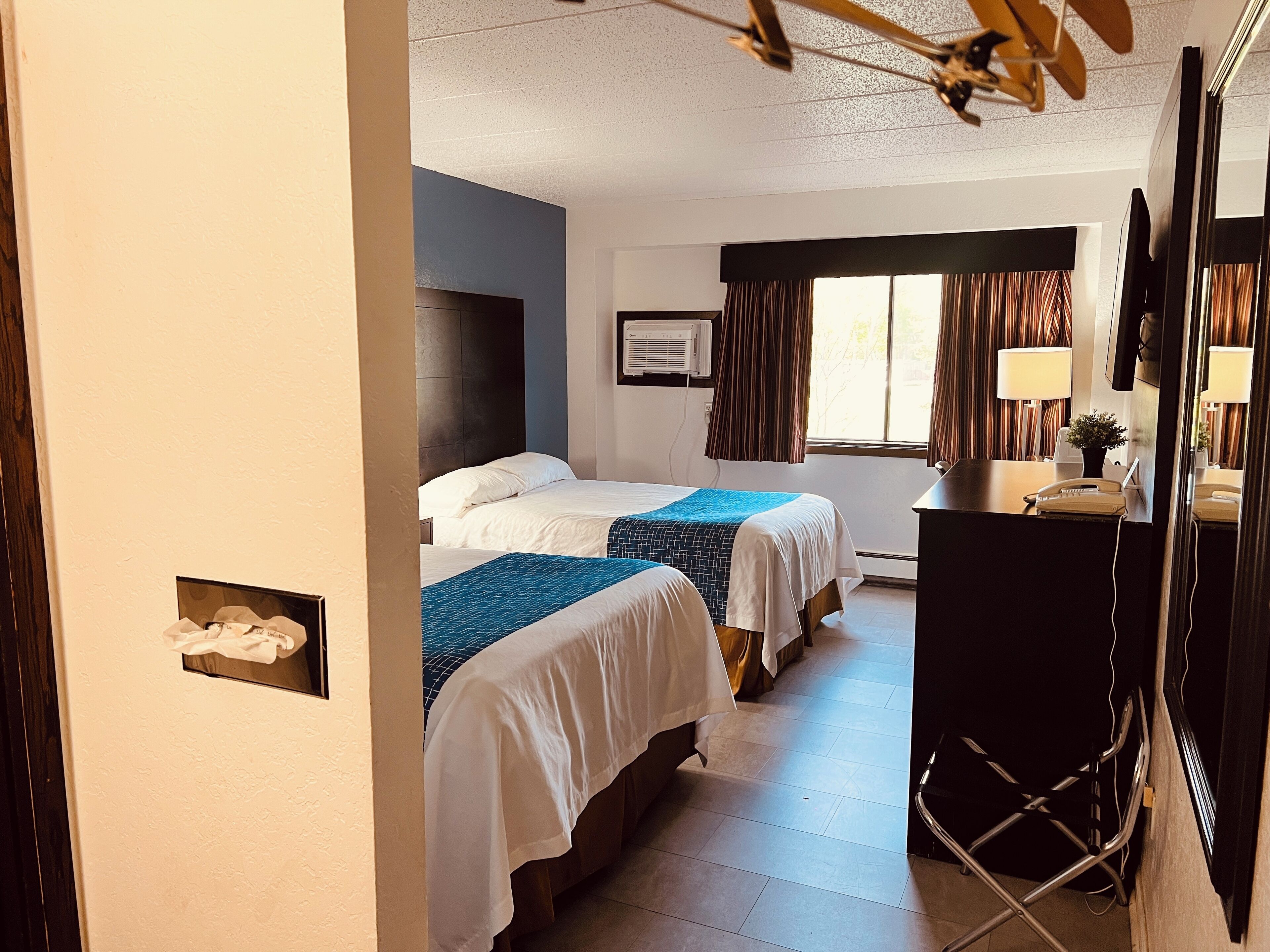 A hotel room with two beds, a desk with a telephone, a mirror, and a window with curtains.