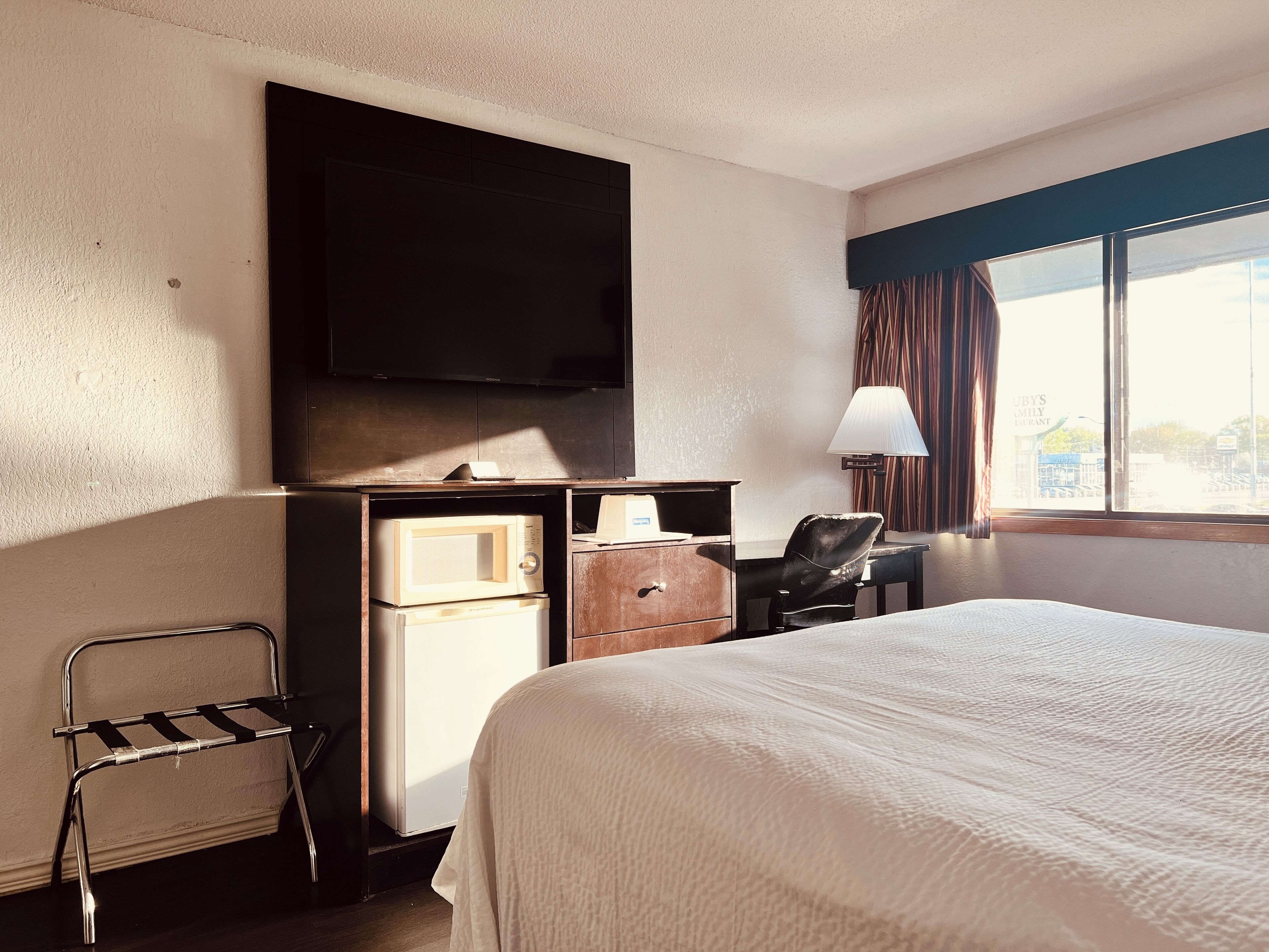 A hotel room with a bed, a desk with a lamp, a chair, a television mounted on the wall, and a window with curtains.