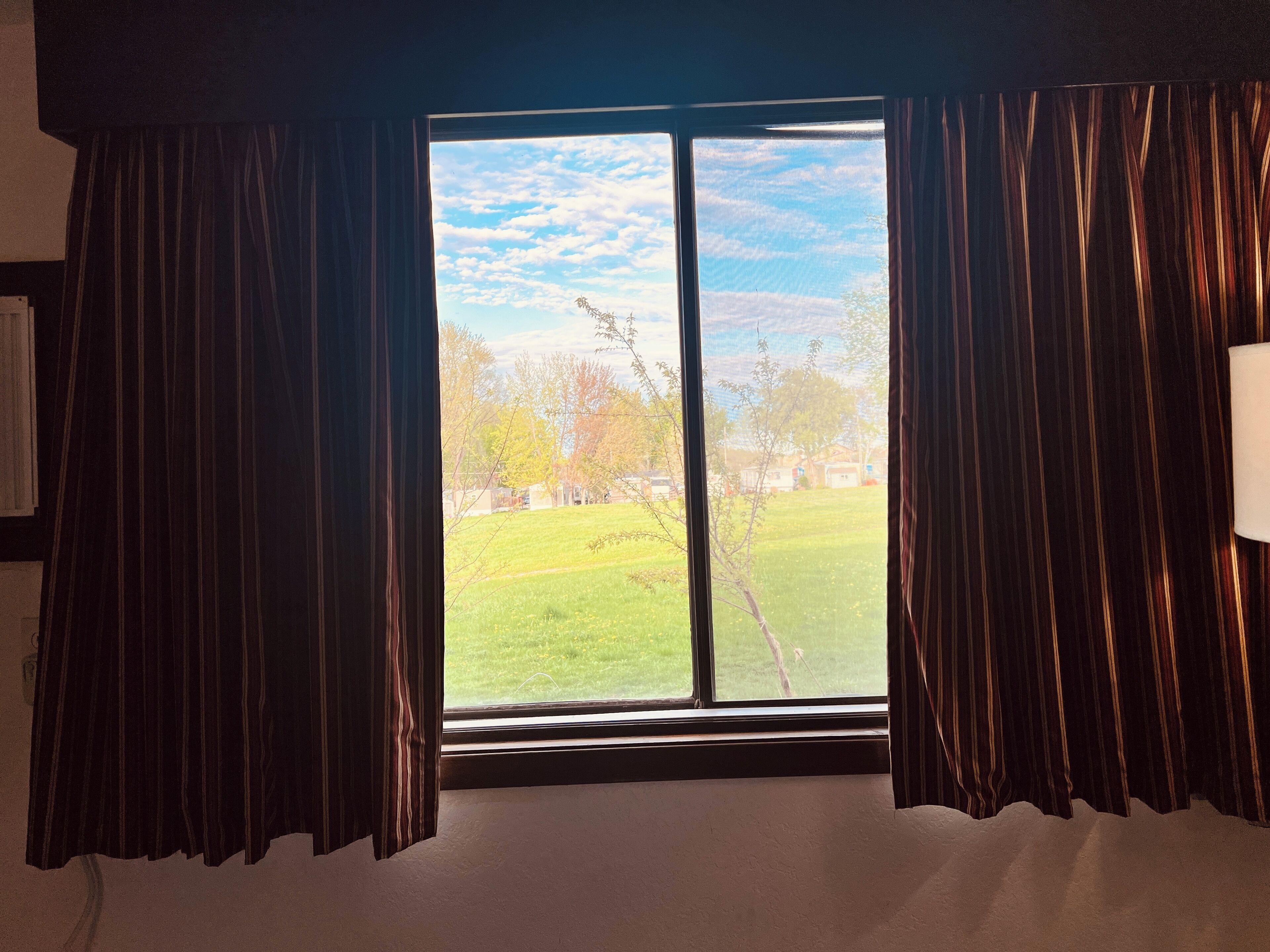 Una habitación con un ventanal grande, cortinas y vistas a un área verde con árboles y casas.