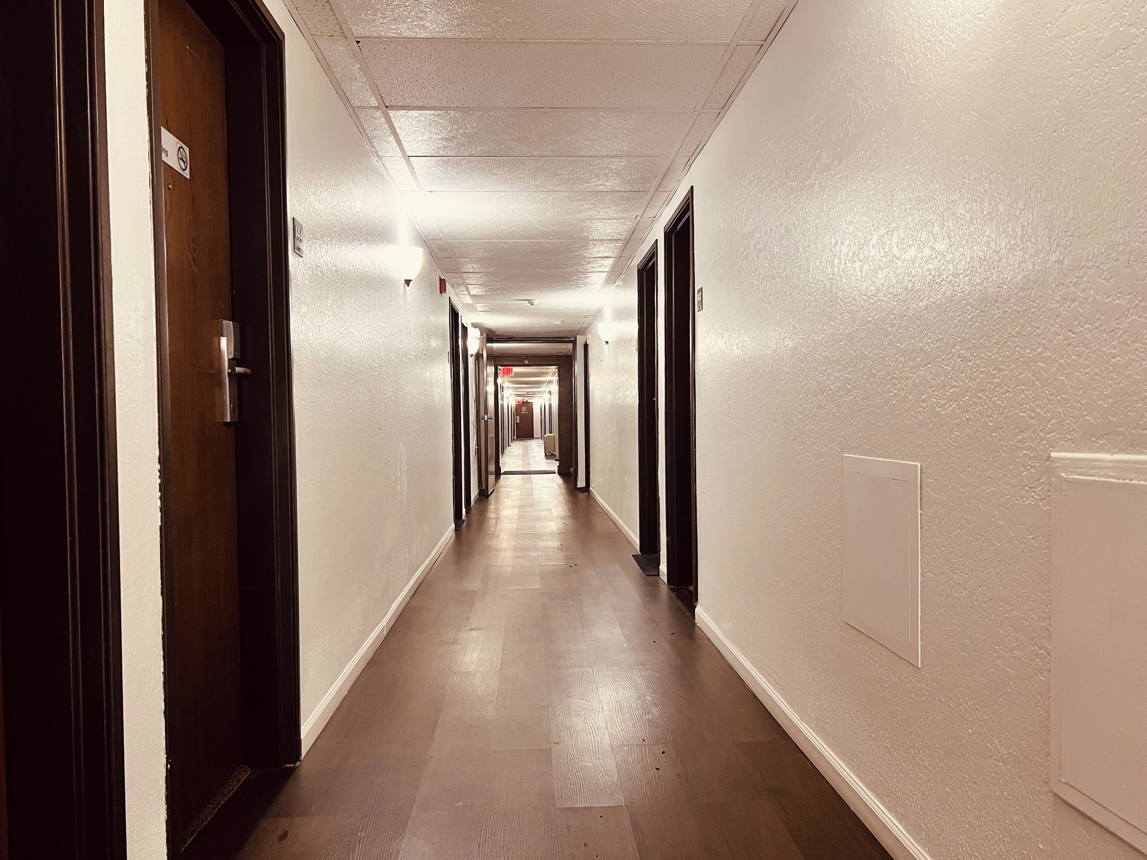 Un pasillo largo y estrecho con pisos de madera y paredes blancas, con puertas a ambos lados y una señal de salida visible.