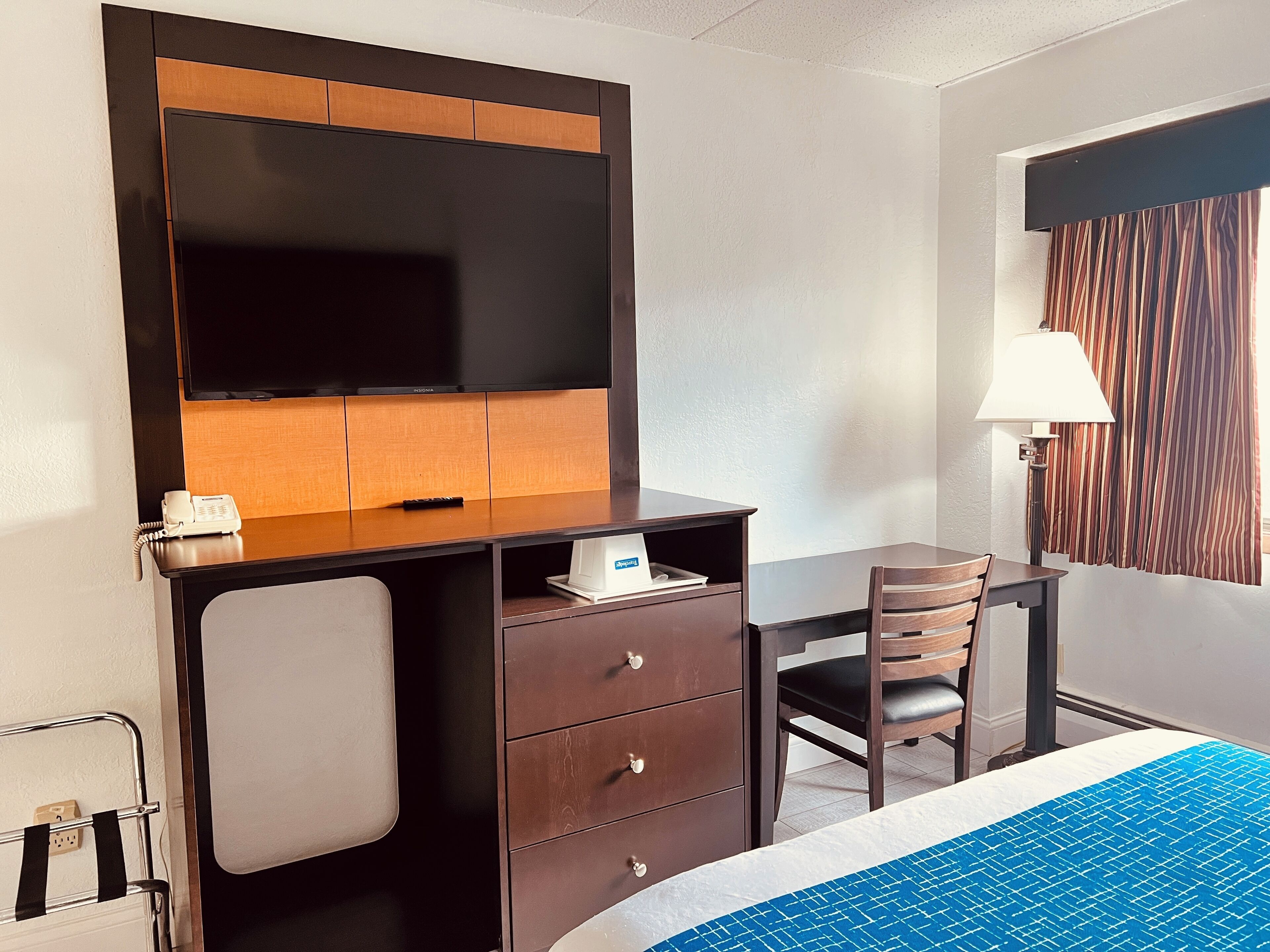 A hotel room with a desk, chair, and bed. There is a television on the wall, a lamp, and a telephone.