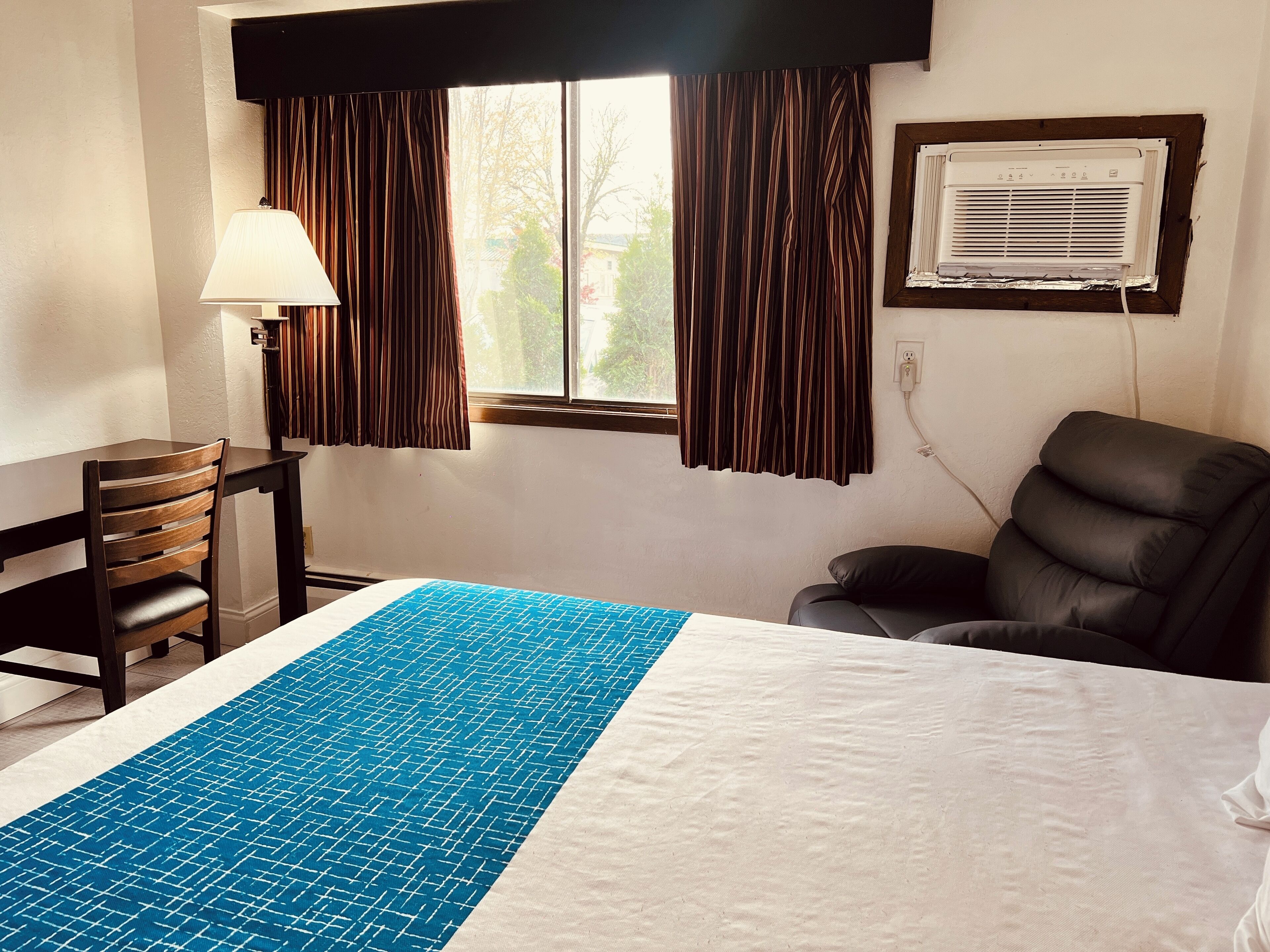 A hotel room with a bed, a desk, a chair, a window with curtains, and an air conditioning unit.