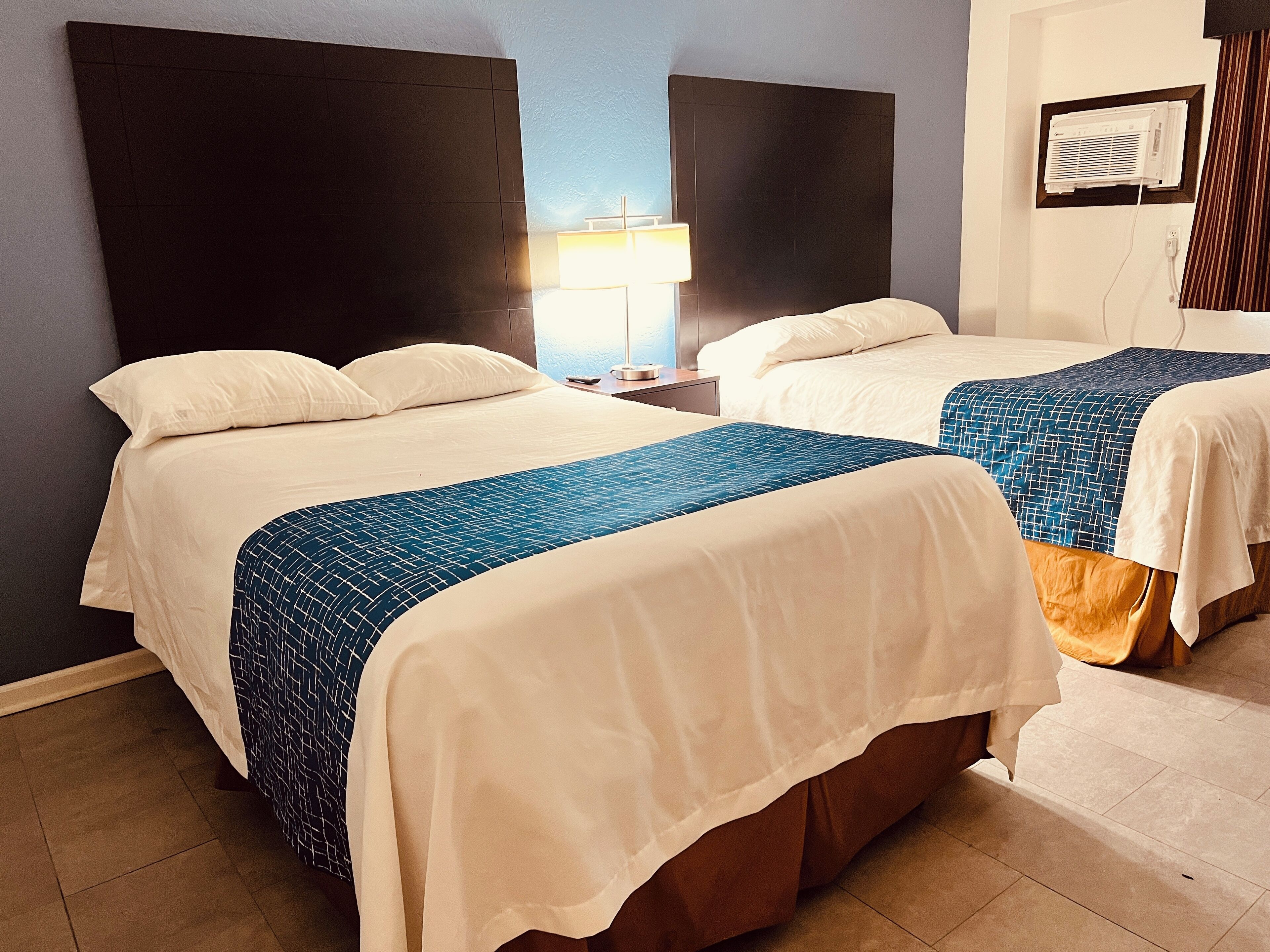 A hotel room with two beds, a nightstand, a lamp, and a wall-mounted air conditioner.