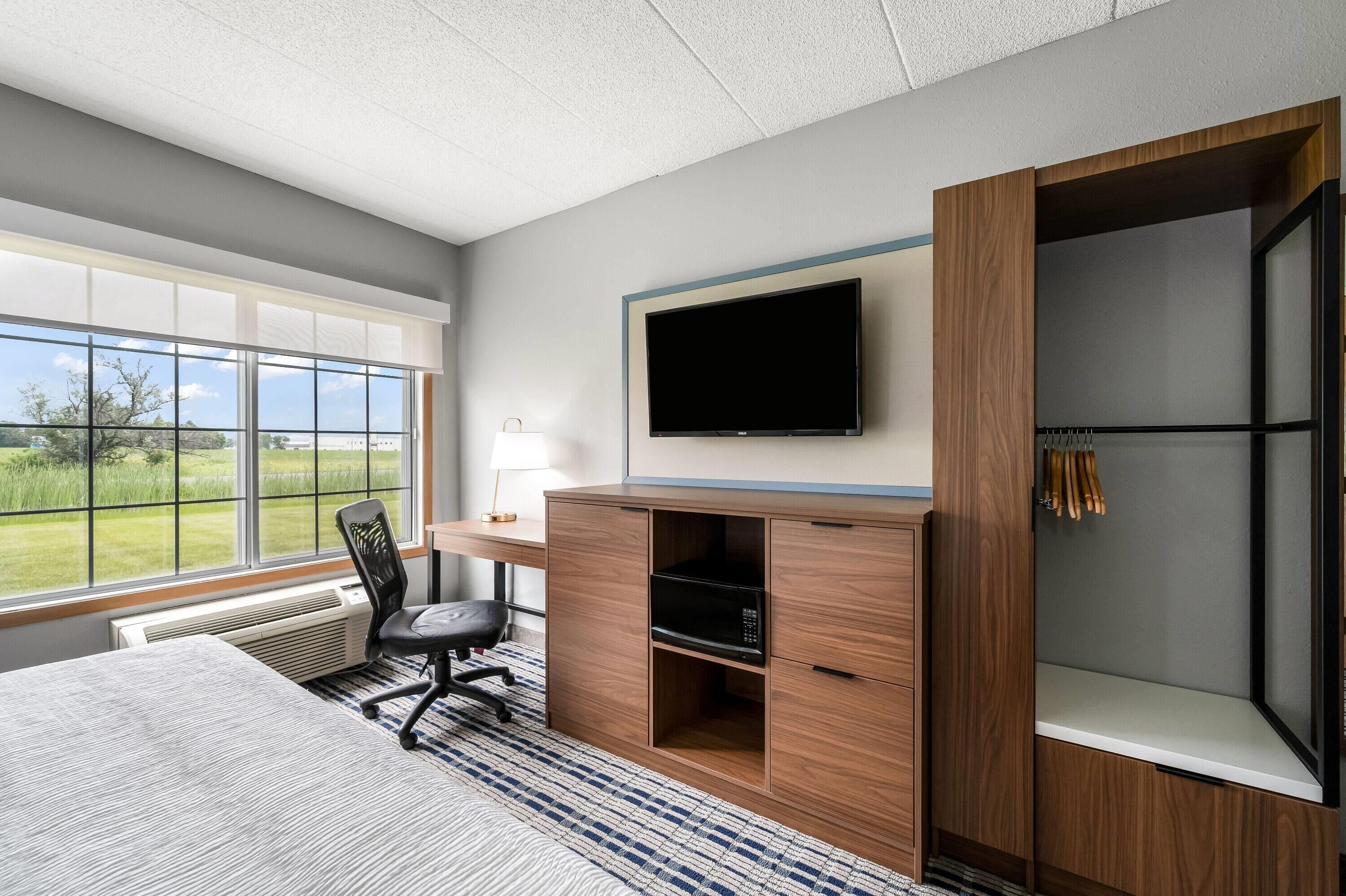 A hotel room with a large window, a flat-screen TV, a desk, a chair, and a wardrobe.
