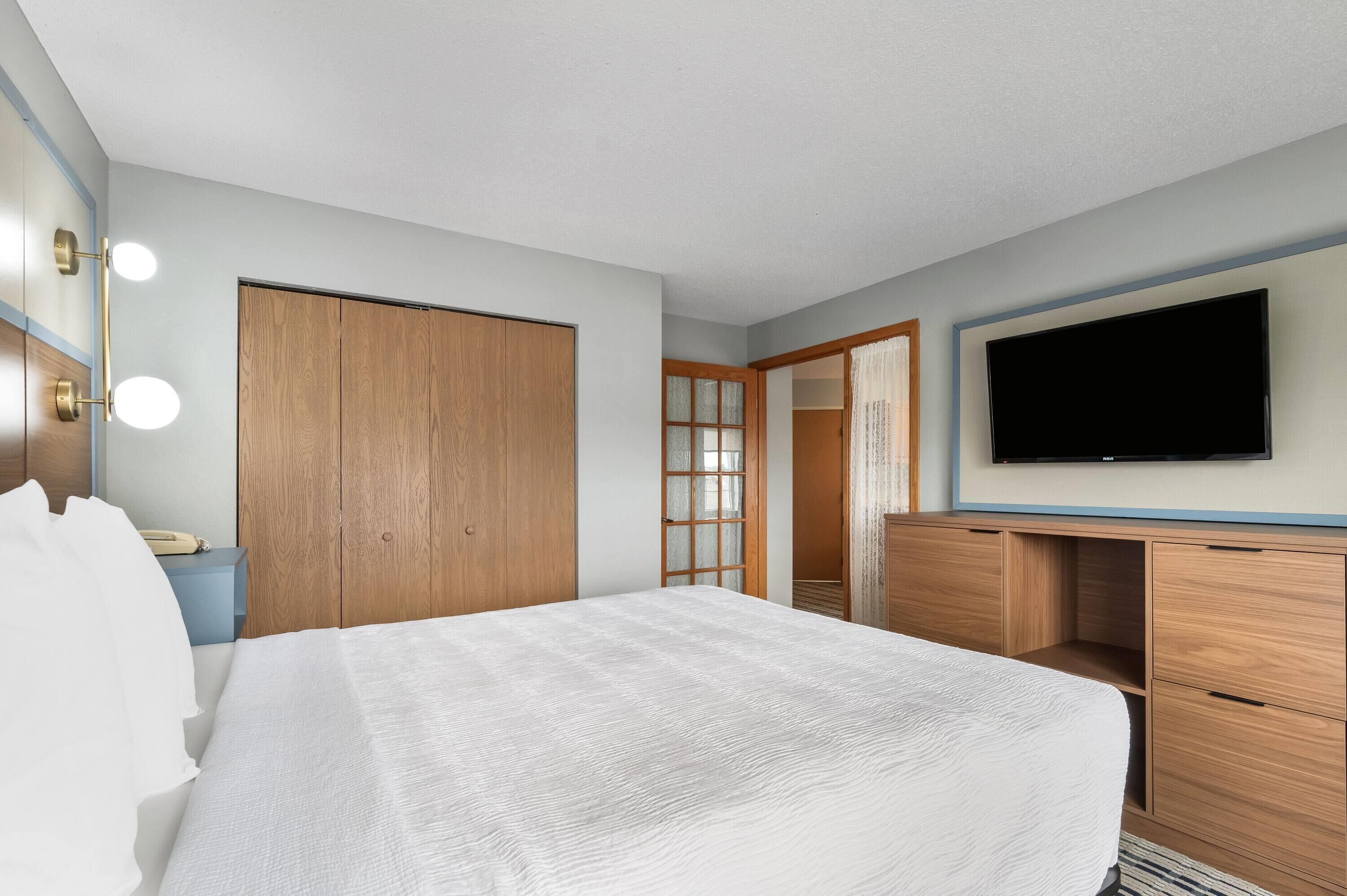A hotel room with a large bed, wooden wardrobe, a desk with drawers, a flat-screen TV, and a mirror with lights.