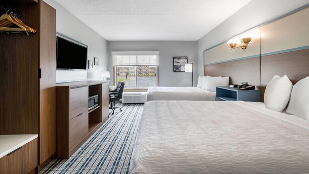 A hotel room with a bed, desk, chair, TV, and a window with a view of buildings. - AmericInn by Wyndham Hutchinson (Hutchinson)