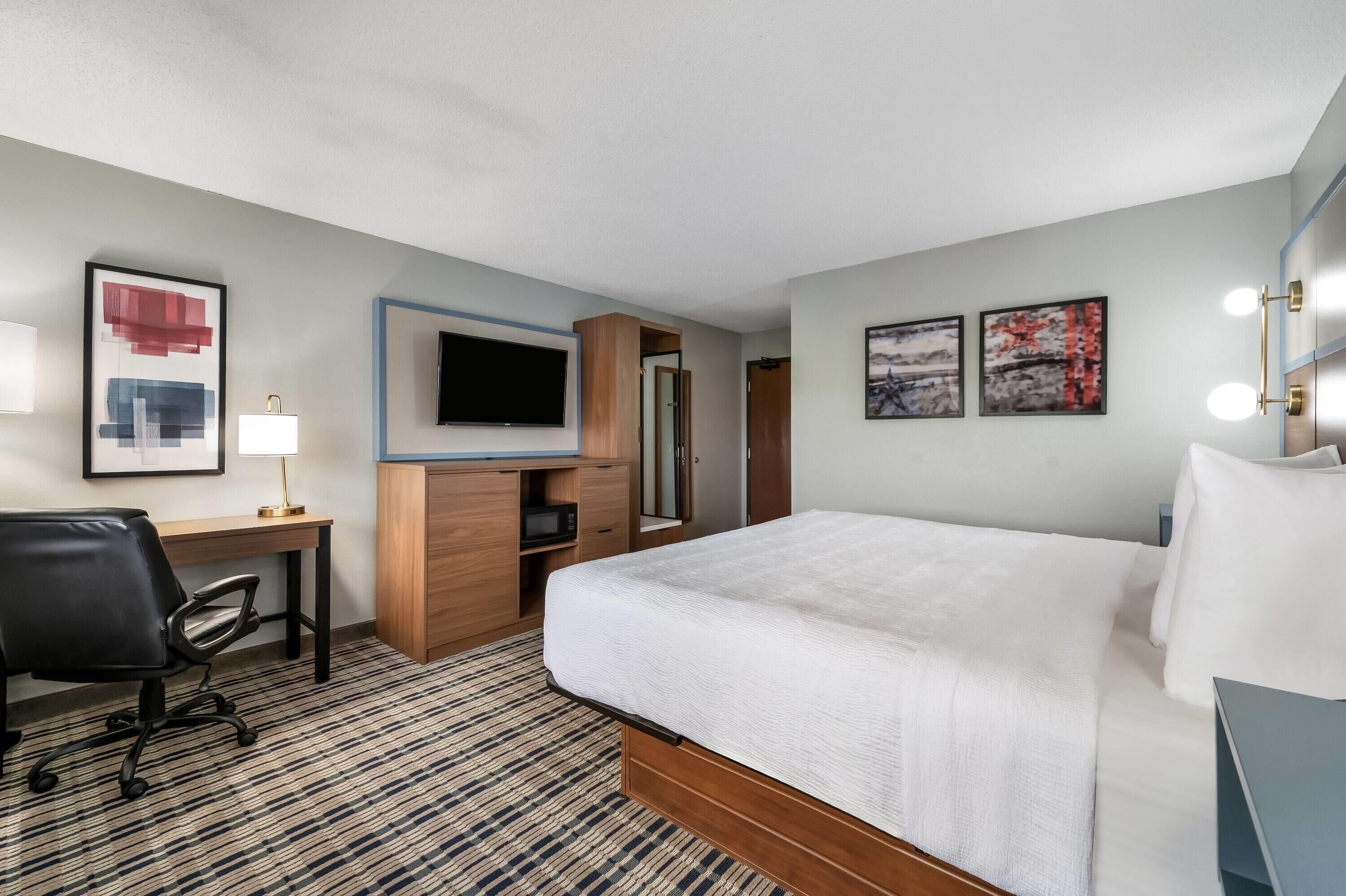 A hotel room with a bed, desk, chair, TV, and artwork.
