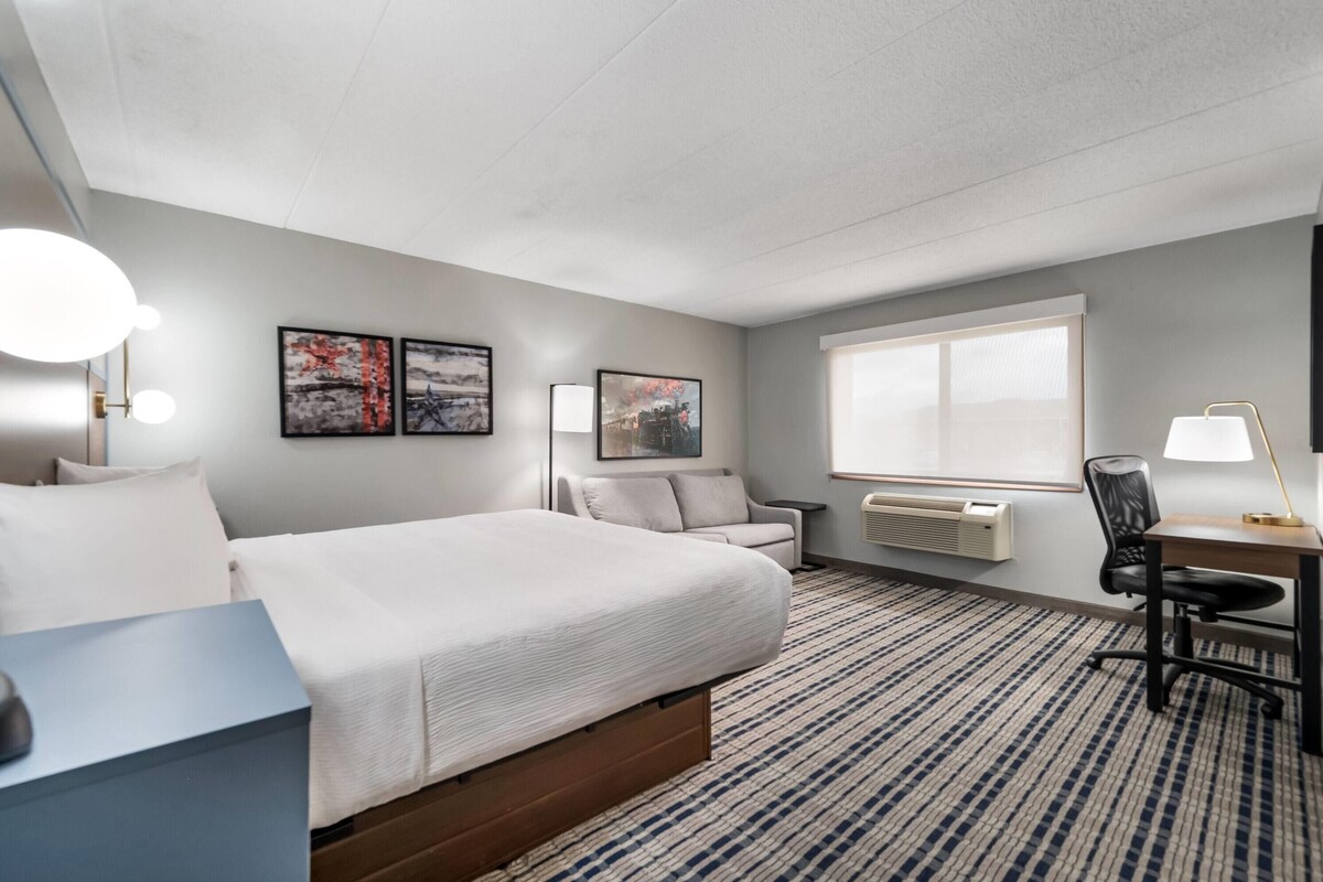 Habitación de hotel con cama, escritorio, silla, lámpara y dos marcos de cuadros en la pared.