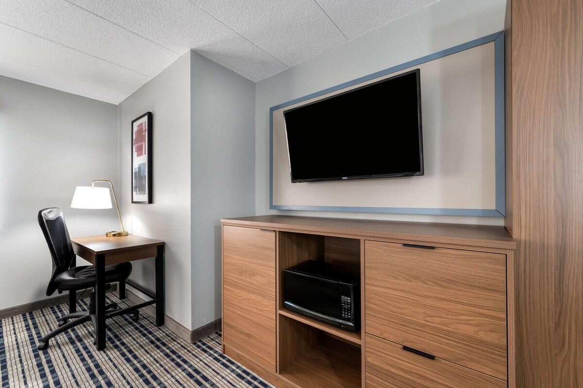 Une chambre d’hôtel équipée d’un téléviseur à écran plat fixé au mur, d’un bureau en bois avec une lampe, d’une chaise et d’un four à micro-ondes placé dans un meuble en bois.