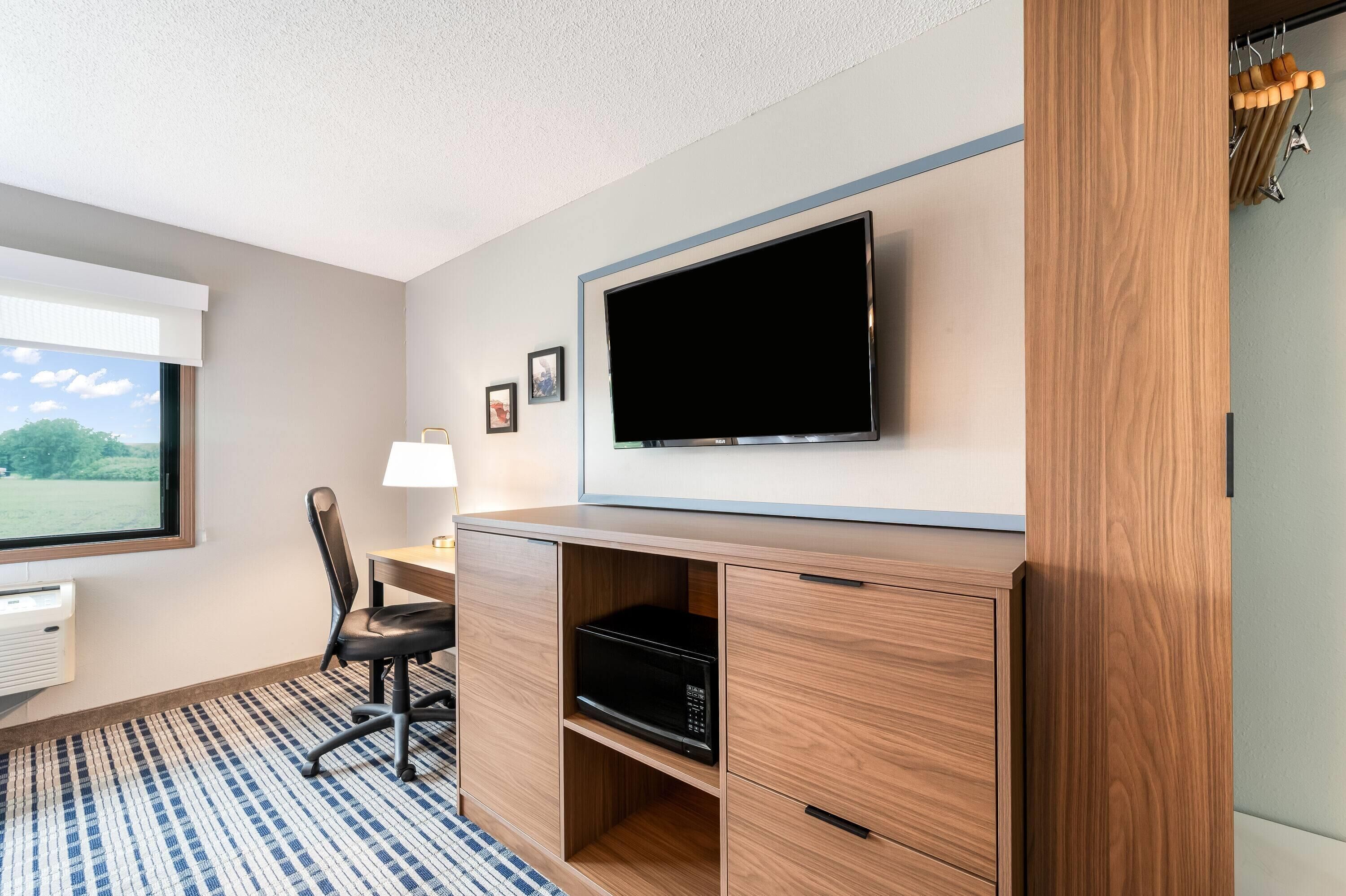 A room with a flat-screen TV mounted on the wall, a desk with a lamp, a chair, a microwave, and a view of the outdoors.