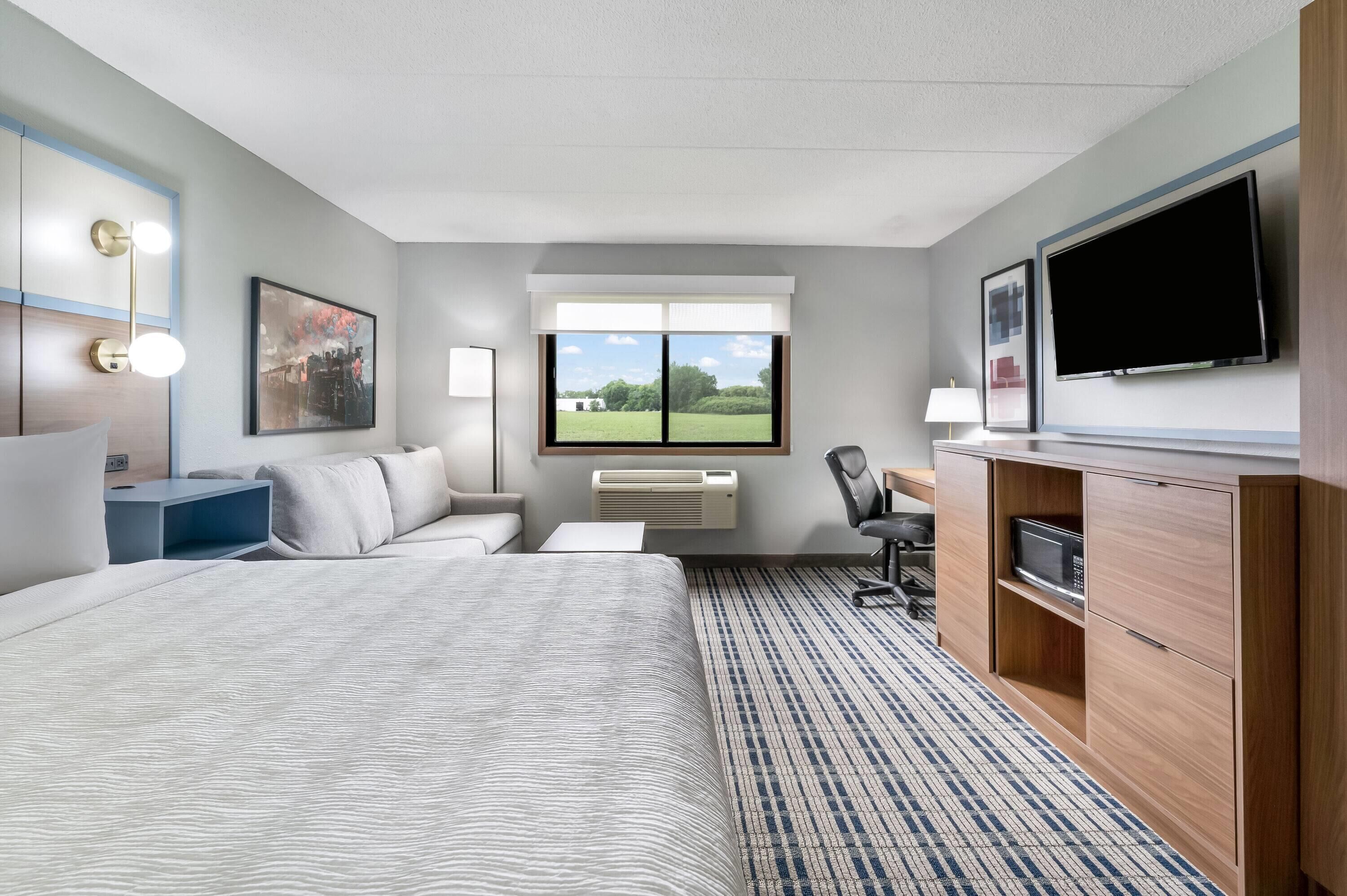 A hotel room with a large bed, a desk, a chair, a flat-screen TV, and a window with a view of a green landscape.
