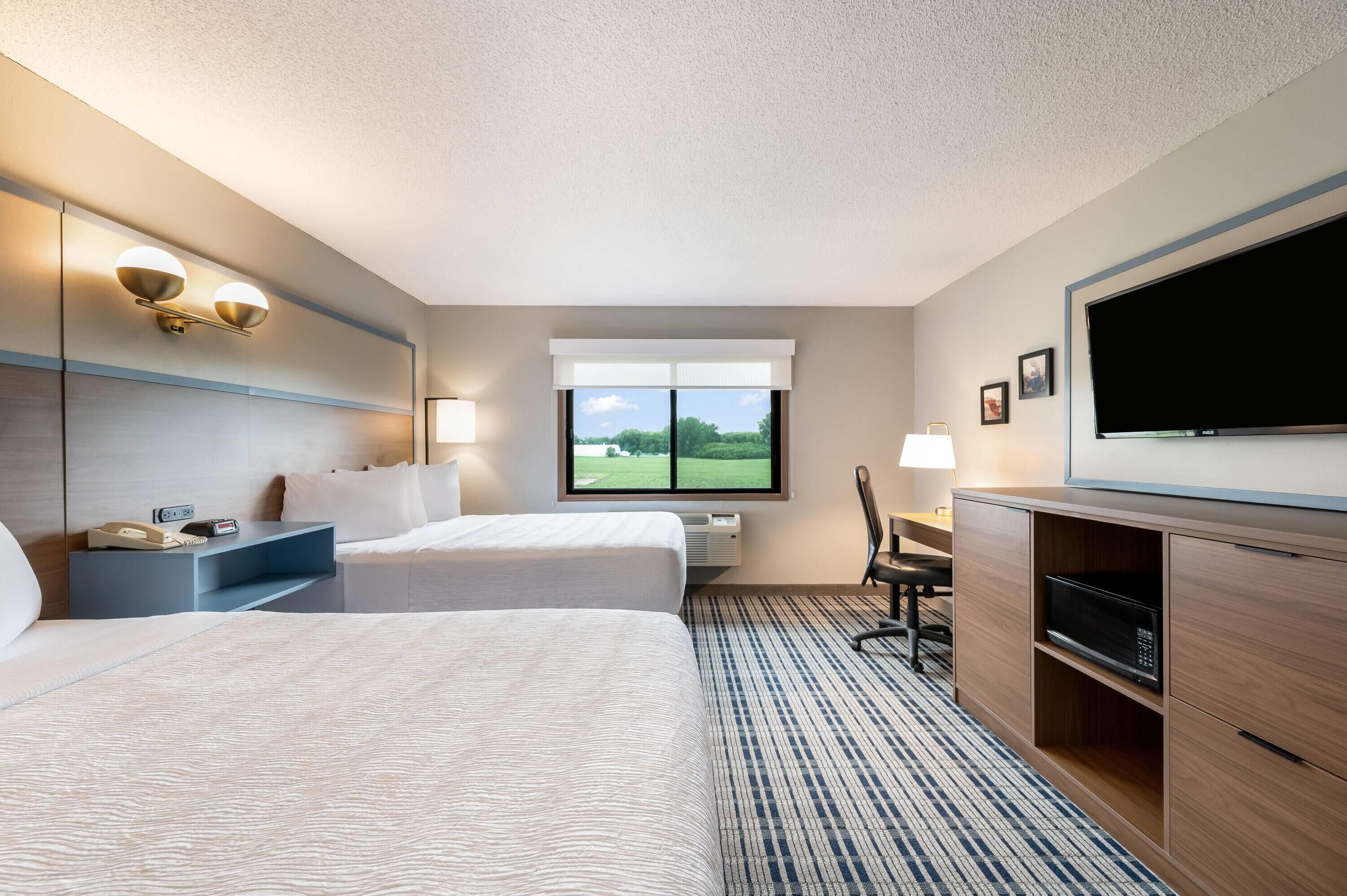 Ein Hotelzimmer mit zwei Betten, einem Schreibtisch, einem Fernseher und einem Fenster mit Blick auf eine grüne Landschaft.
