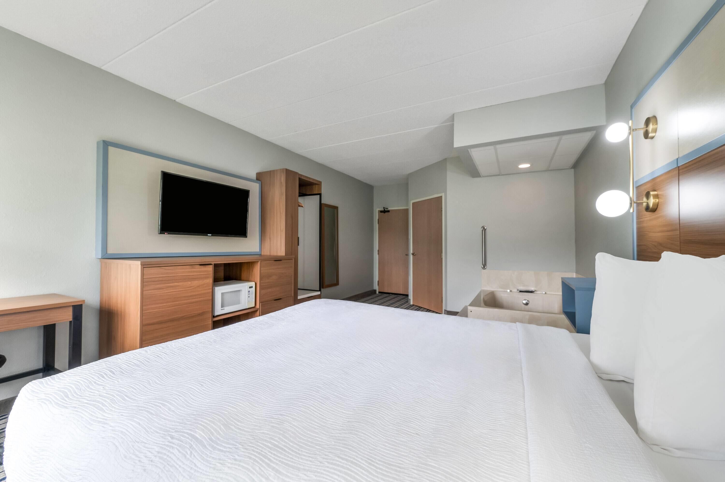 A hotel room with a large bed, a TV mounted on the wall, a wardrobe, a bedside table, and a mirror with lights.