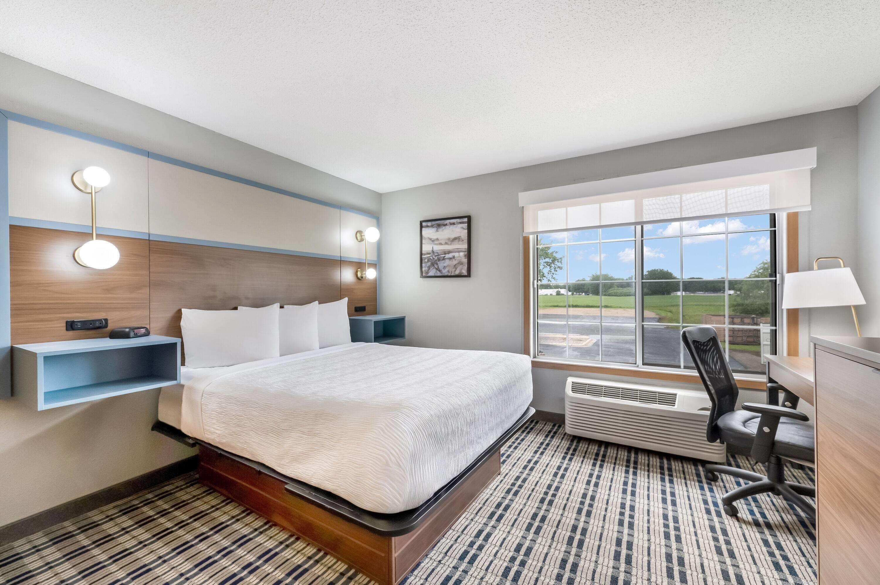 A hotel room with a large bed, bedside tables, a desk with a chair, a window with a view, and a wall-mounted light fixture.