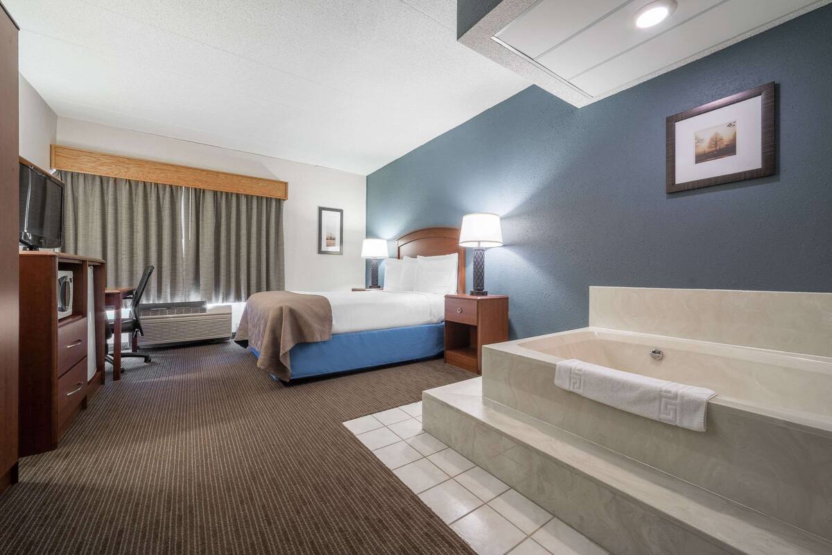Habitación de hotel con cama, lámparas de noche, televisión, escritorio, silla, lámpara de escritorio, ventana con cortinas, un cuadro en la pared y una bañera.