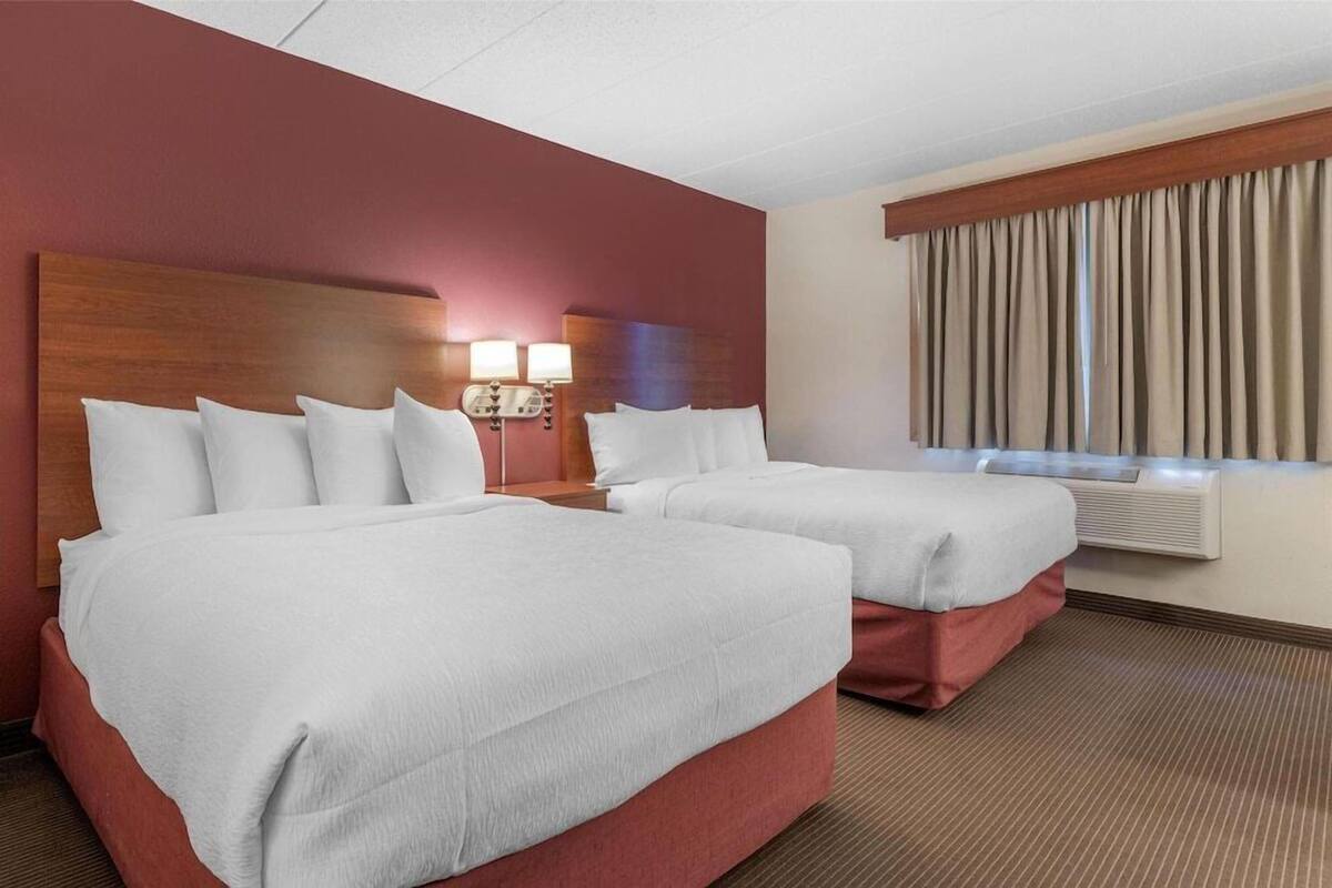A hotel room with two beds, a wooden headboard, a window with curtains, and a wall-mounted air conditioning unit.