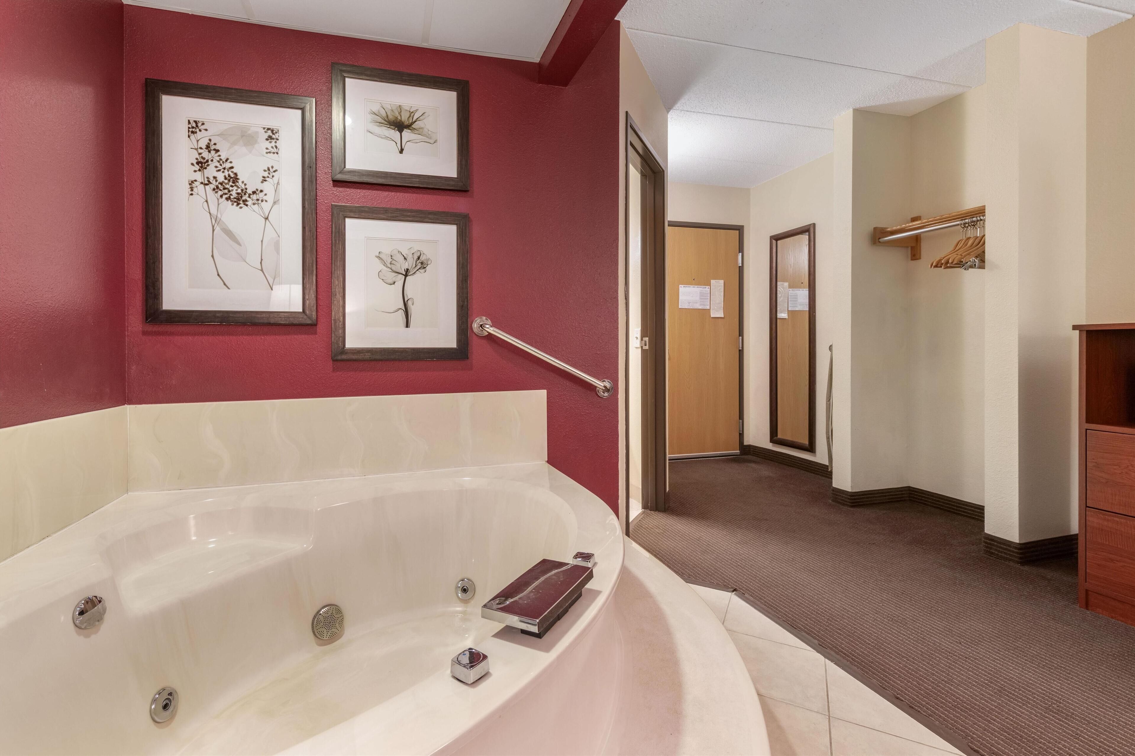 A hotel bathroom with a large bathtub, framed botanical artwork on the wall, a handrail, and a wooden door in the background.