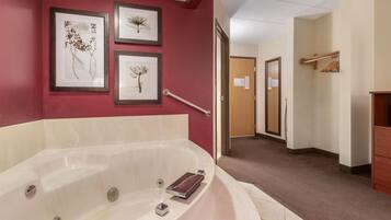 A hotel bathroom with a large bathtub, framed botanical artwork on the wall, a handrail, and a wooden door in the background.