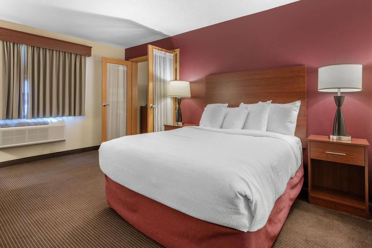 A hotel room with a large bed, a wooden headboard, two bedside tables with lamps, a window with curtains, and a wall-mounted air conditioning unit.