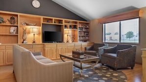A cozy living room with a sofa, armchairs, a coffee table, and a TV. There are wooden shelves with books and decorative items, and a large window with blinds.
