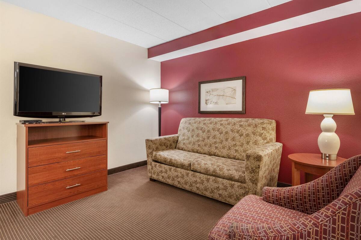 A hotel room with a flat-screen TV on a wooden stand, a floral-patterned sofa, a red accent wall, and a small table with a lamp.