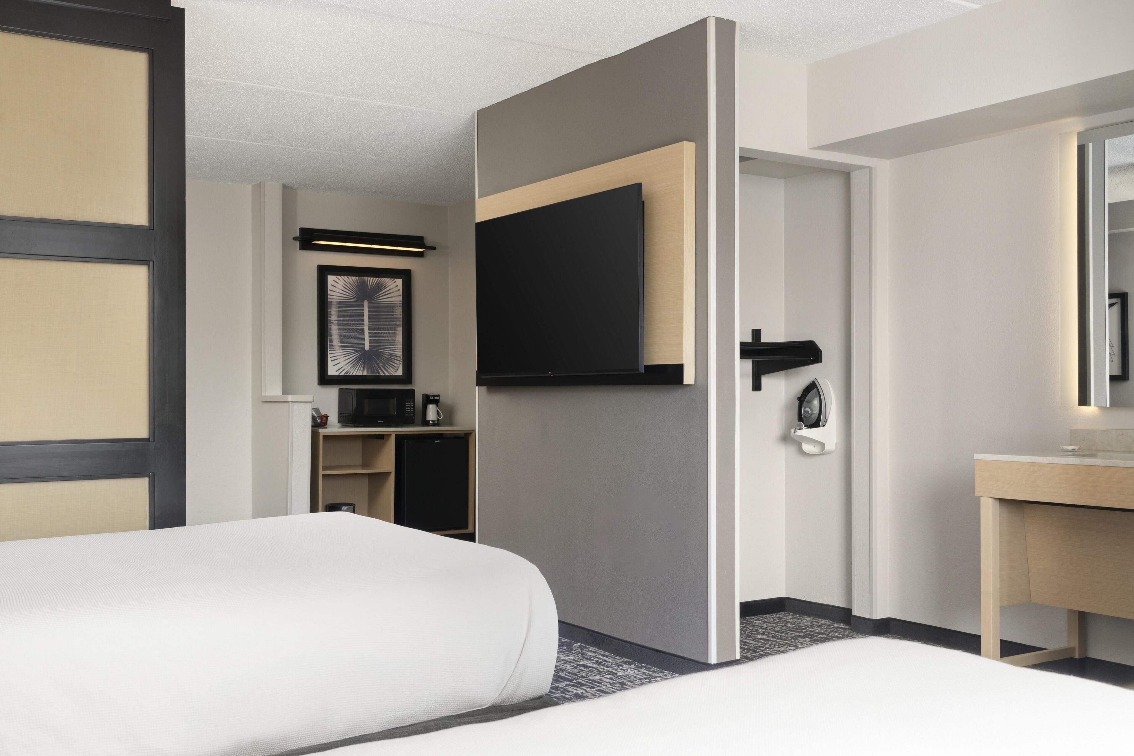 Habitación de hotel con dos camas, un televisor montado en la pared, un escritorio con espejo y una pequeña zona de kitchenette.