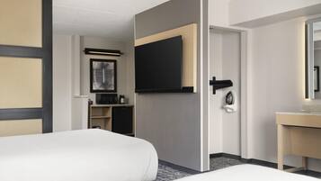 Habitación de hotel con dos camas, un televisor montado en la pared, un escritorio con espejo y una pequeña zona de kitchenette.