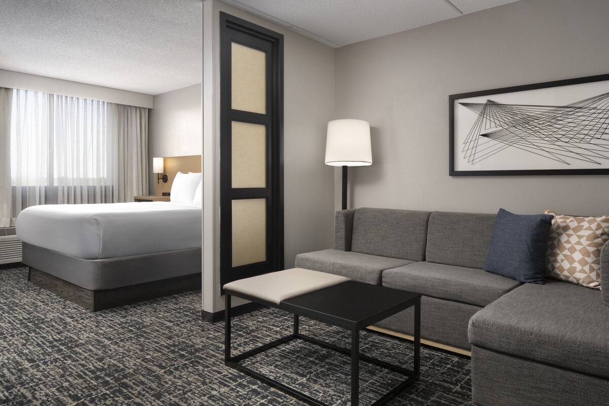 A hotel room with a bed, a sofa, a small table, a lamp, and a framed abstract artwork on the wall.