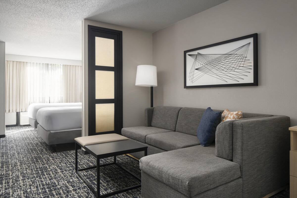 A hotel room with a gray sofa, a small table, a lamp, a framed abstract artwork, and a bed with white linens.