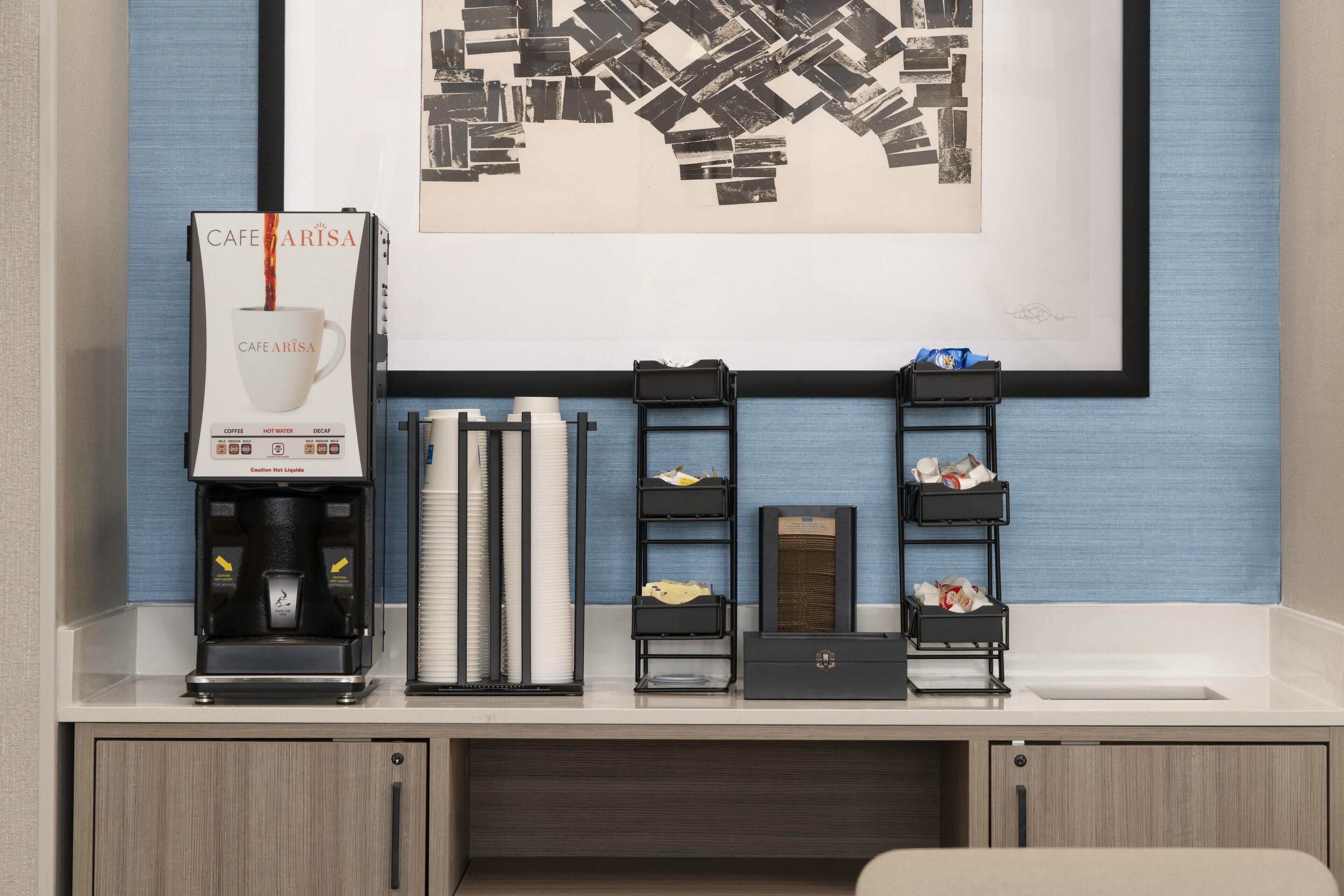 Een koffiehoek met een koffieapparaat met branding, een stapel papieren bekers en een ingelijste afbeelding aan de muur.