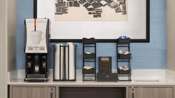 Een koffiehoek met een koffieapparaat met branding, een stapel papieren bekers en een ingelijste afbeelding aan de muur.