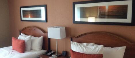A hotel room with two beds, each with white bedding and red pillows, a bedside table with a lamp, and framed artwork on the walls.