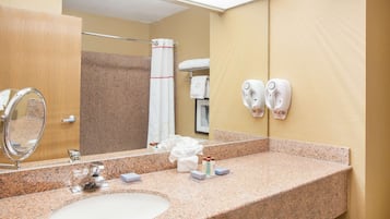 Ein Hotelbadezimmer mit Waschbecken auf Granitplatte, einem großen Spiegel, einem Handtuchhalter und zwei an der Wand befestigten Haartrocknern.