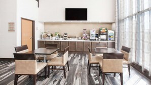 Un comedor de hotel con una larga mesa y sillas, una encimera de cocina con electrodomésticos y un amplio ventanal con cortinas.