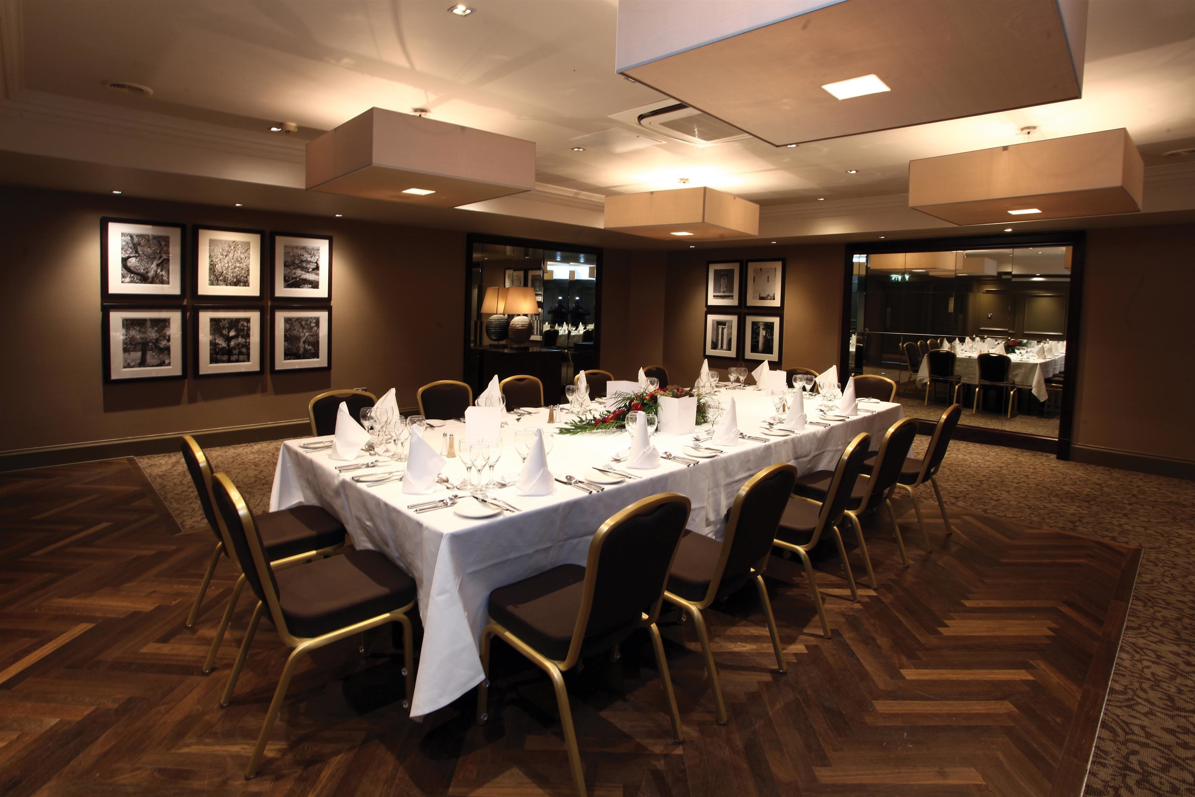 A conference room with a long table set for a meeting, surrounded by chairs, with framed pictures on the walls and a view into another room.