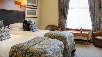 A hotel room with two beds, a small table with a bottle and glass, a chair, and a window with curtains.