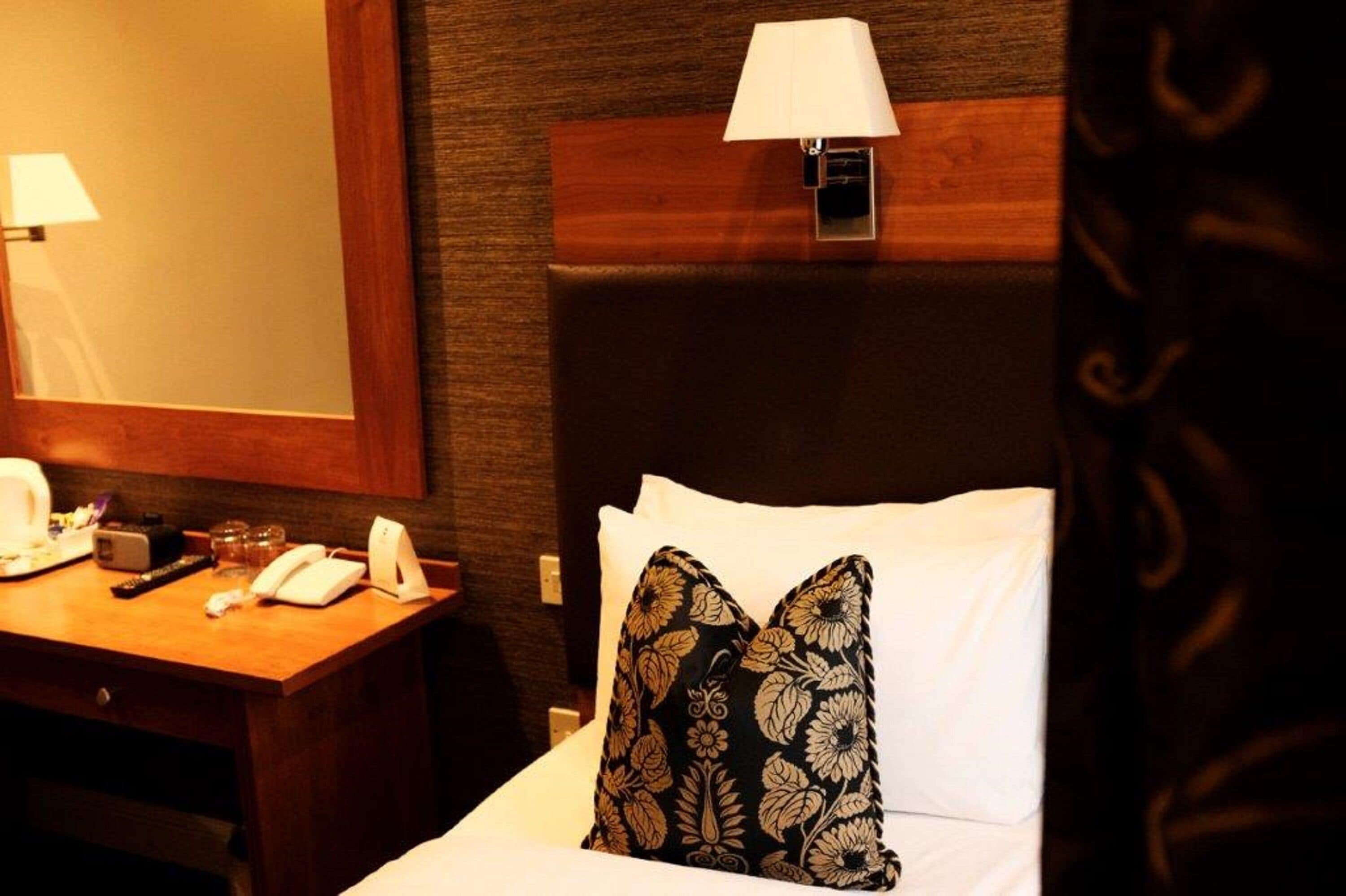 Habitación de hotel con cabecera de madera, lámpara de noche, teléfono y almohada decorativa.