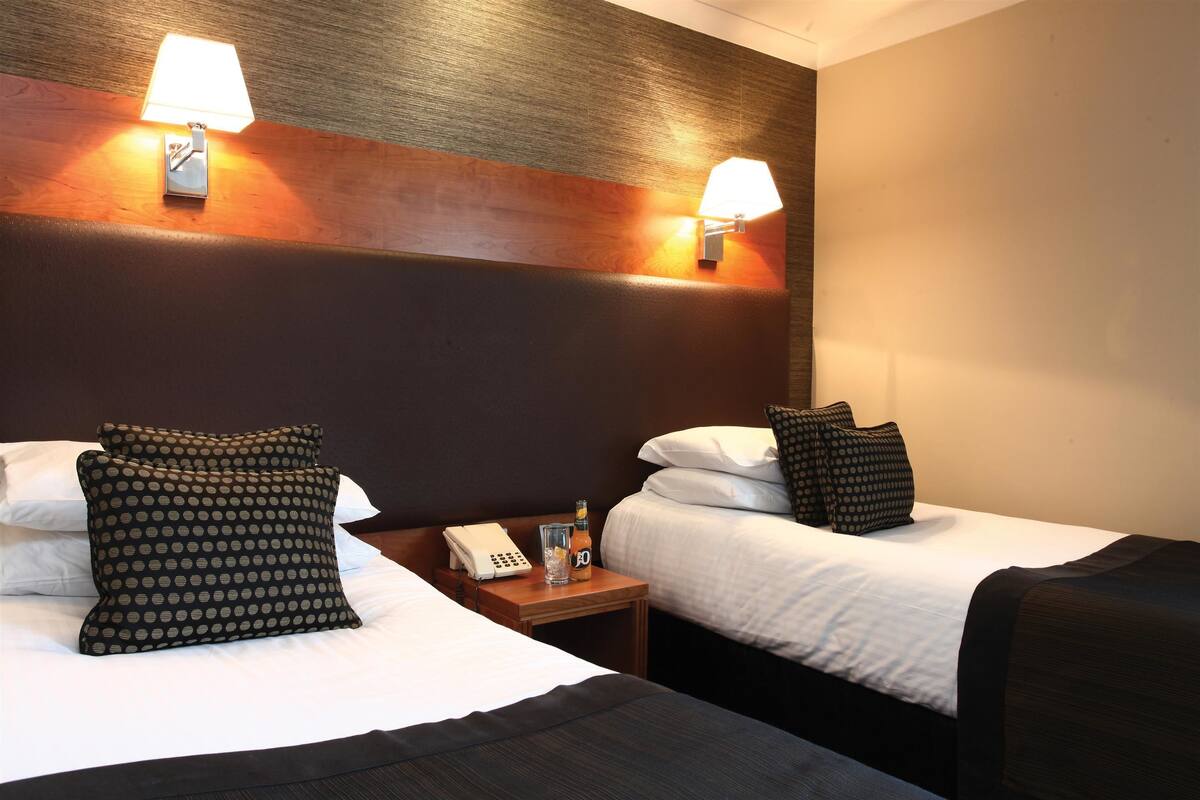 A hotel room with two beds, each with a pillow and a lamp. There is a nightstand with a phone and a glass of water.