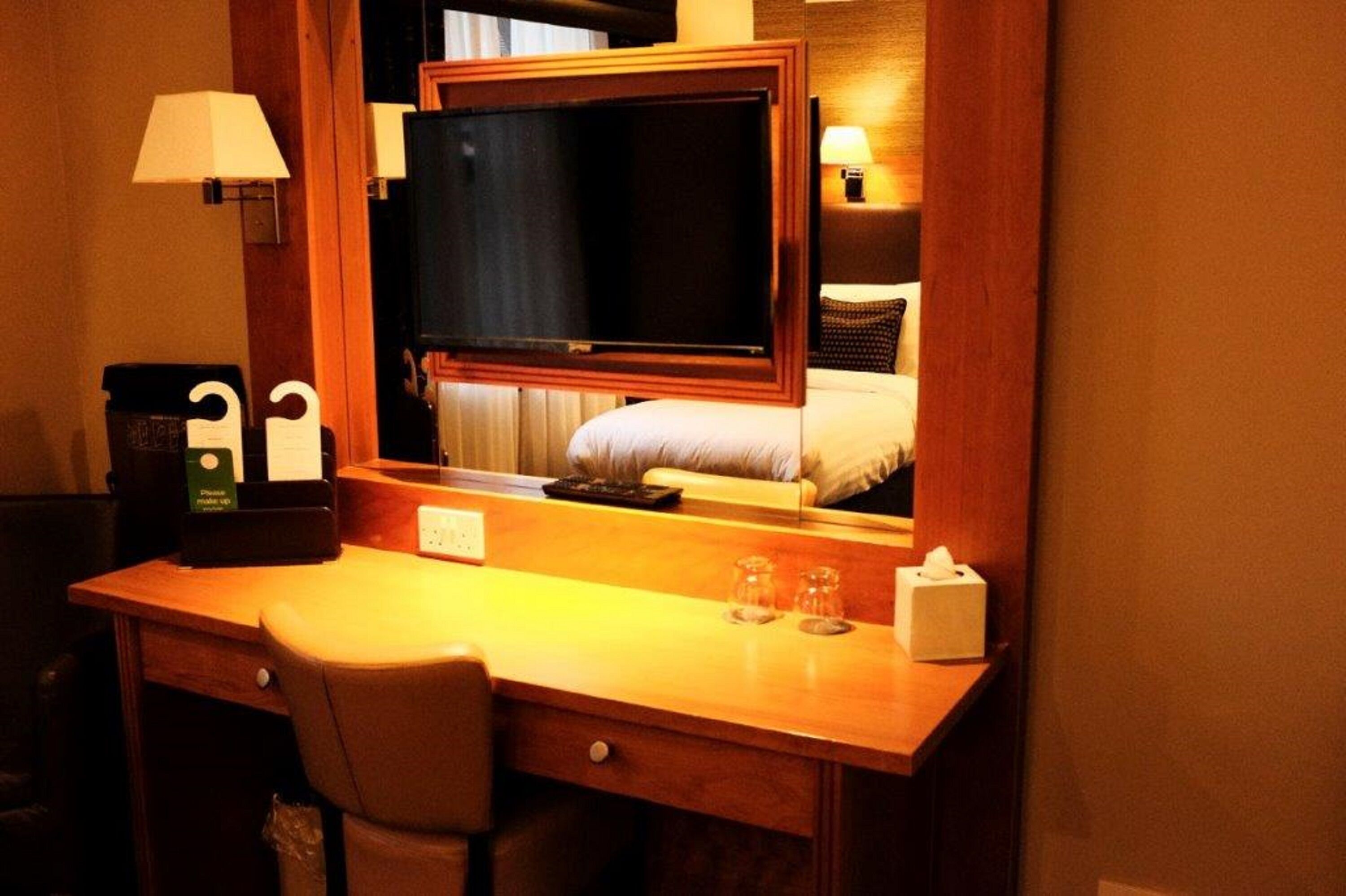 Une chambre d’hôtel avec un bureau en bois, une télévision, un lit recouvert d’une couette blanche, une table de chevet avec une lampe, et une petite table sur laquelle se trouvent une boîte de mouchoirs et des verres.
