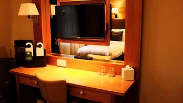 Une chambre d’hôtel avec un bureau en bois, une télévision, un lit recouvert d’une couette blanche, une table de chevet avec une lampe, et une petite table sur laquelle se trouvent une boîte de mouchoirs et des verres.