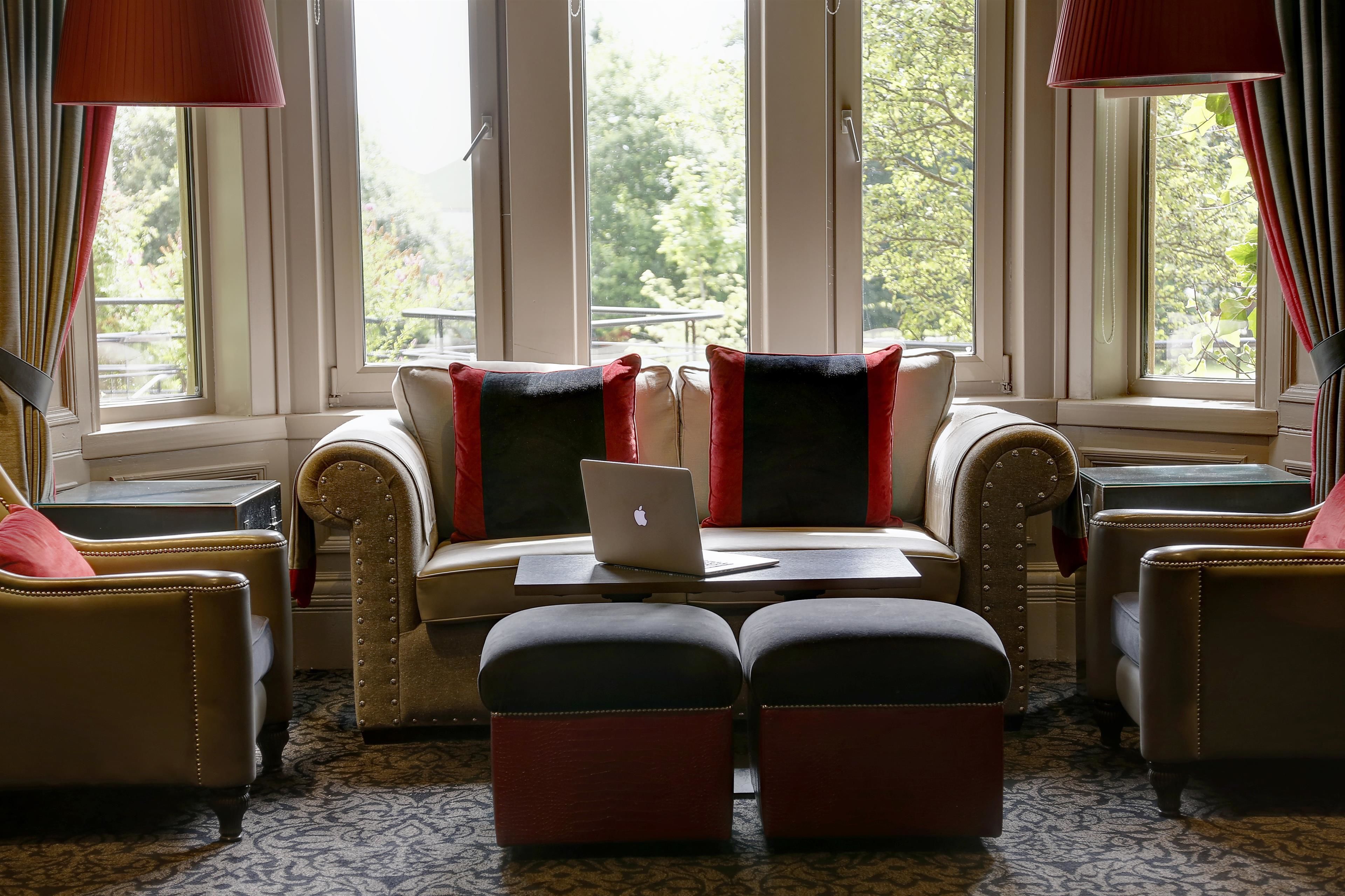 Un coin salon confortable avec un canapé et des poufs, un ordinateur portable posé sur une petite table, et de grandes fenêtres avec des rideaux.