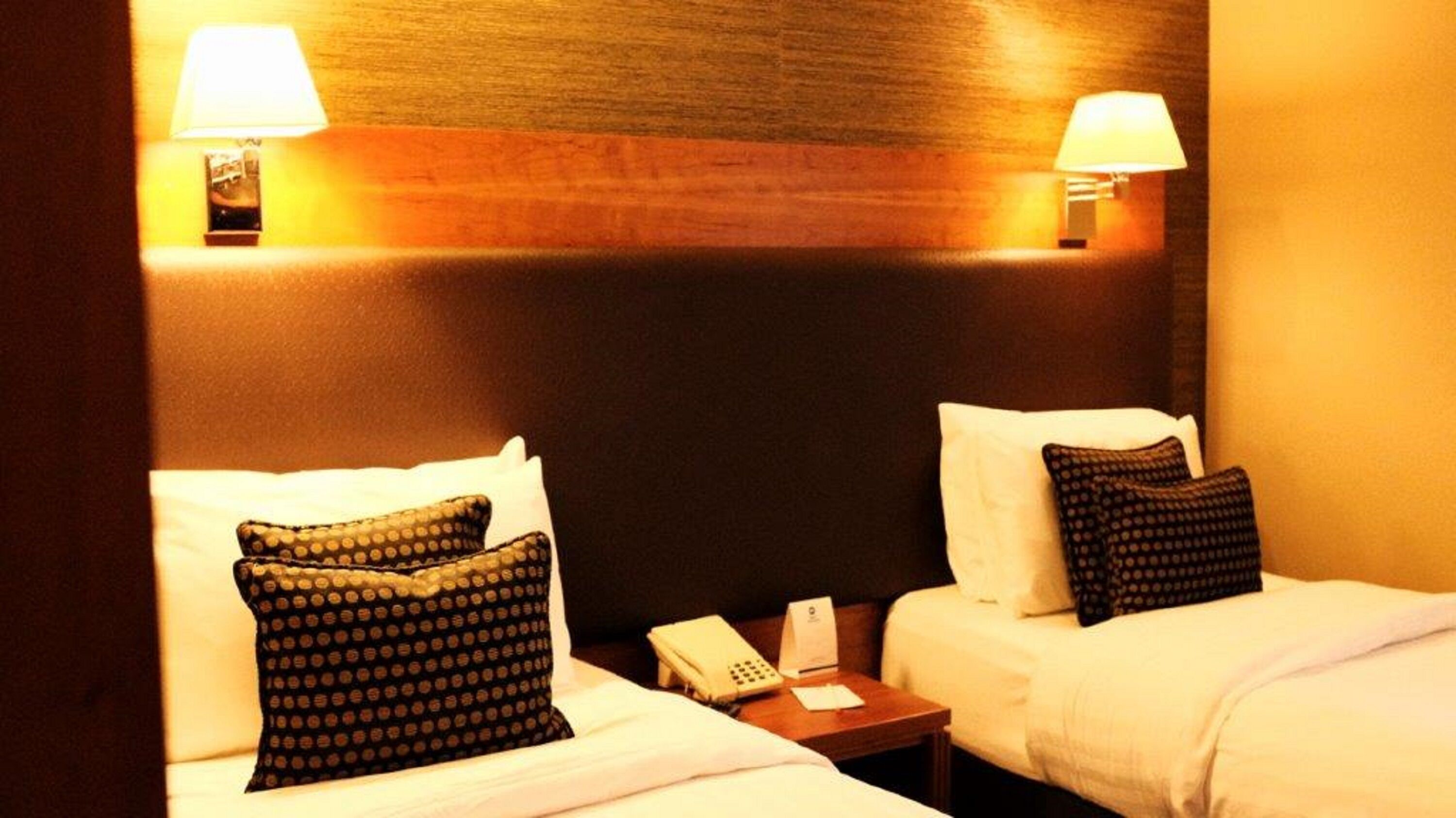 Una camera d'albergo con due letti, ciascuno con un cuscino e un comodino. Sul comodino c'è un telefono e a parete una lampada.