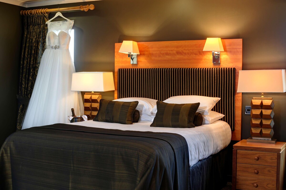 Una habitación de hotel con una cama grande, dos lámparas de noche, un cabecero de madera y un vestido de novia colgado.