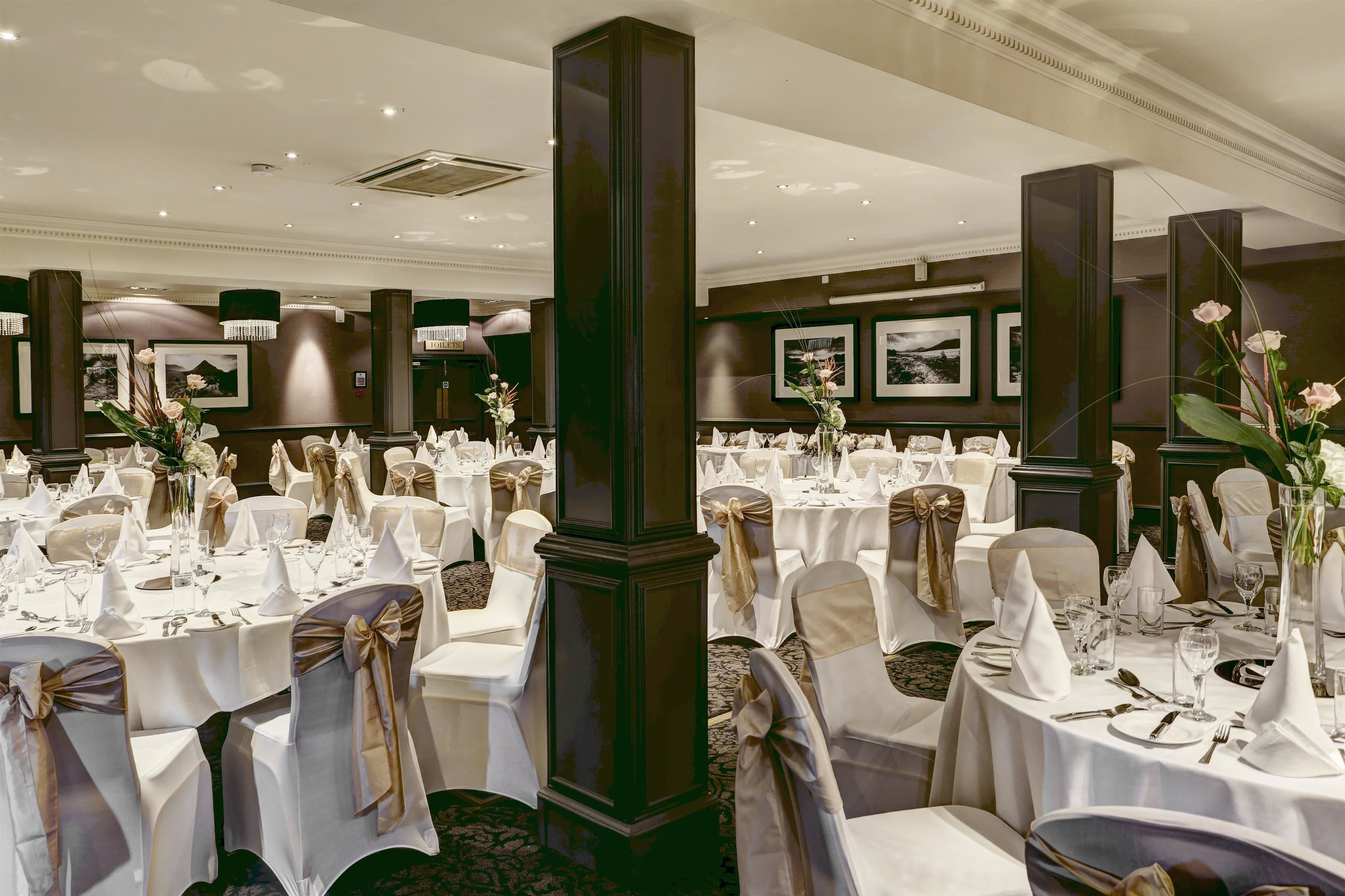 Un salón de banquetes con mesas redondas dispuestas para un evento formal, con manteles blancos, fajas doradas y centros de mesa florales.