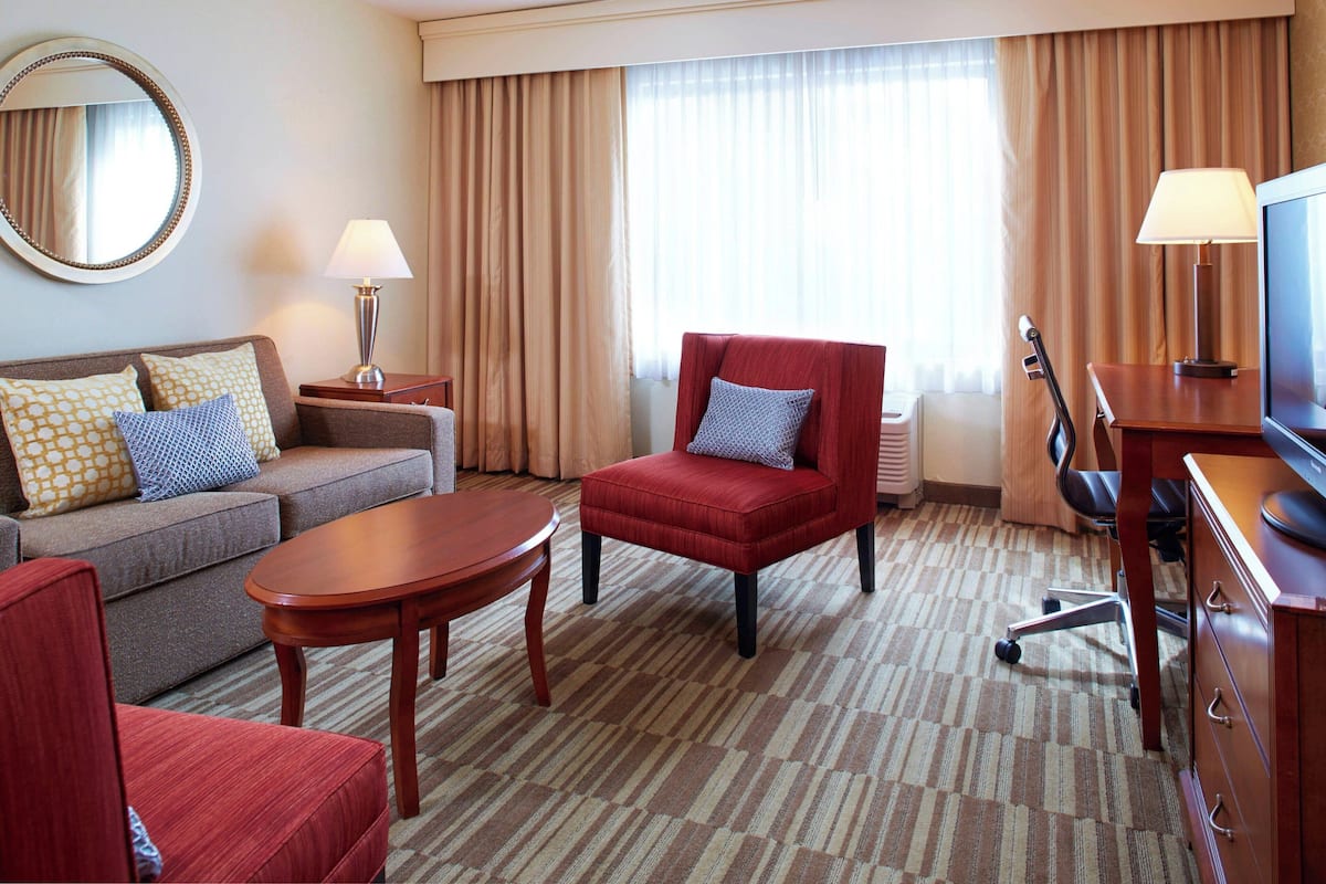 A hotel room with a sofa, armchair, TV, lamp, and a large window with curtains.