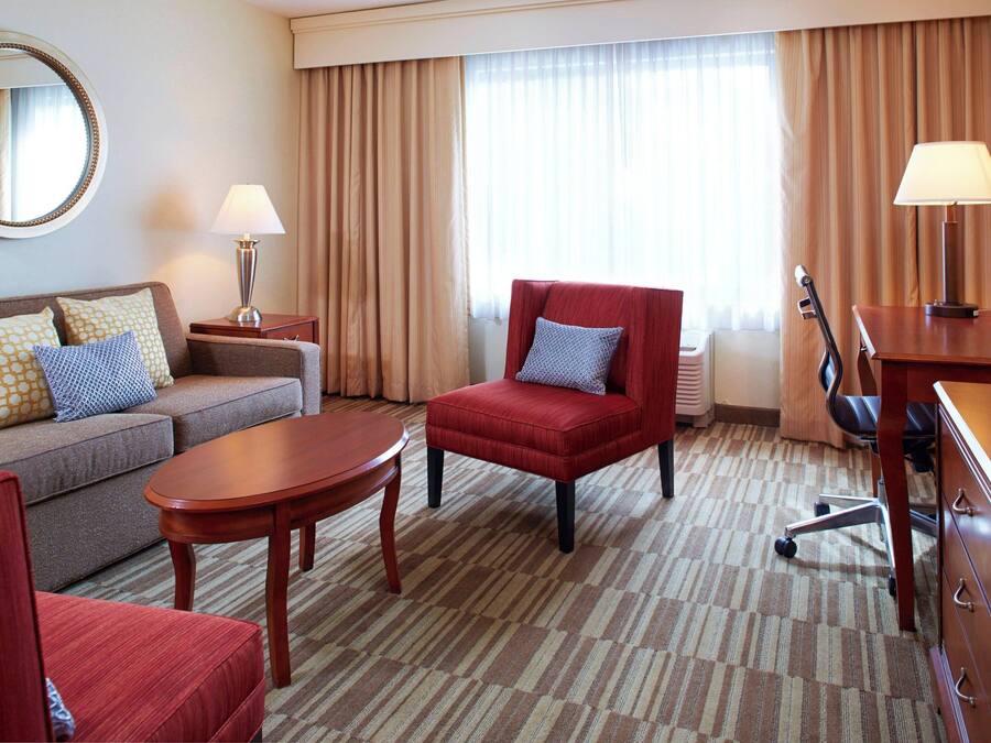 A hotel room with a sofa, armchair, TV, lamp, and a large window with curtains.